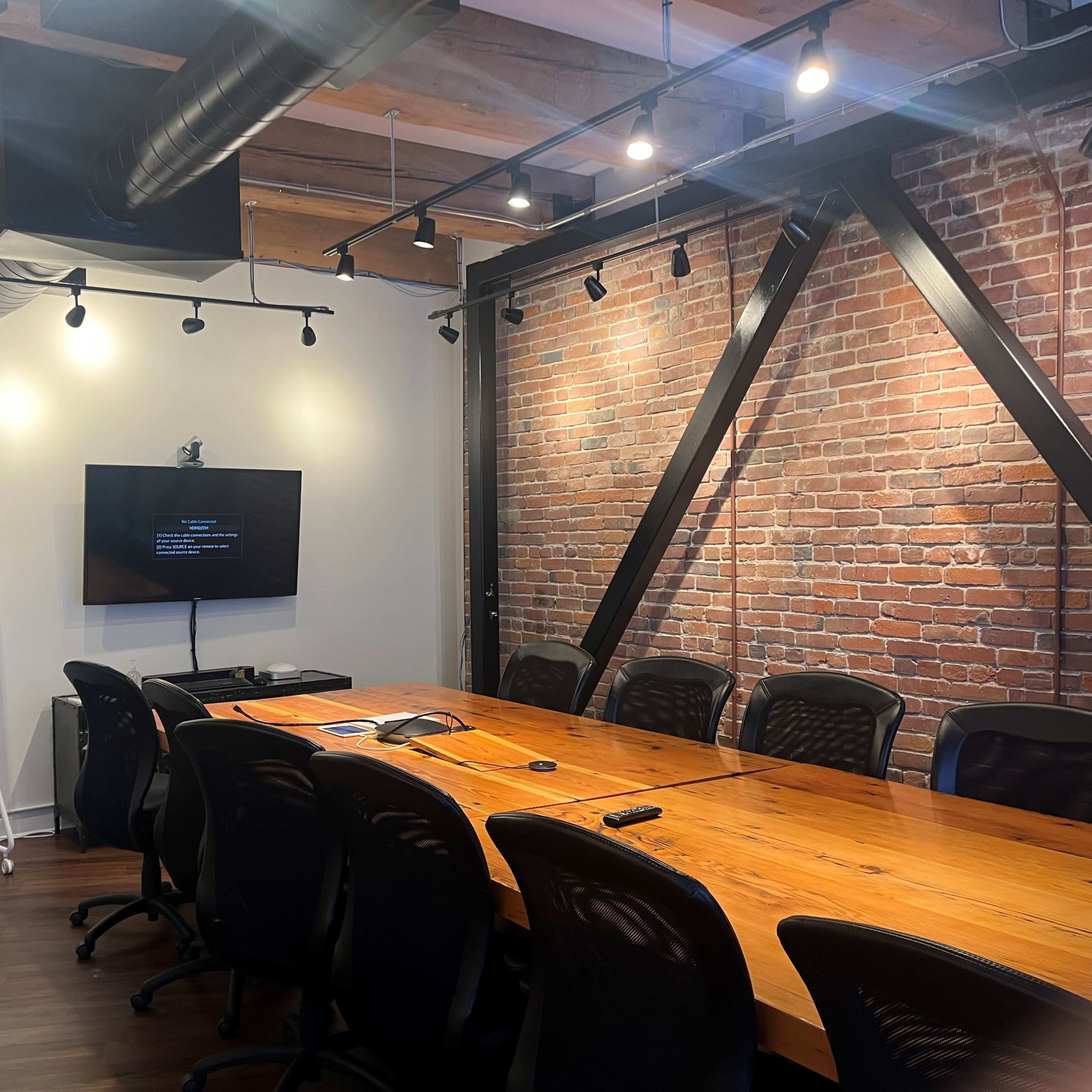 A modern conference room with a long wooden table, black ergonomic chairs, and two wall-mounted screens surrounded by exposed brick walls and overhead lighting.