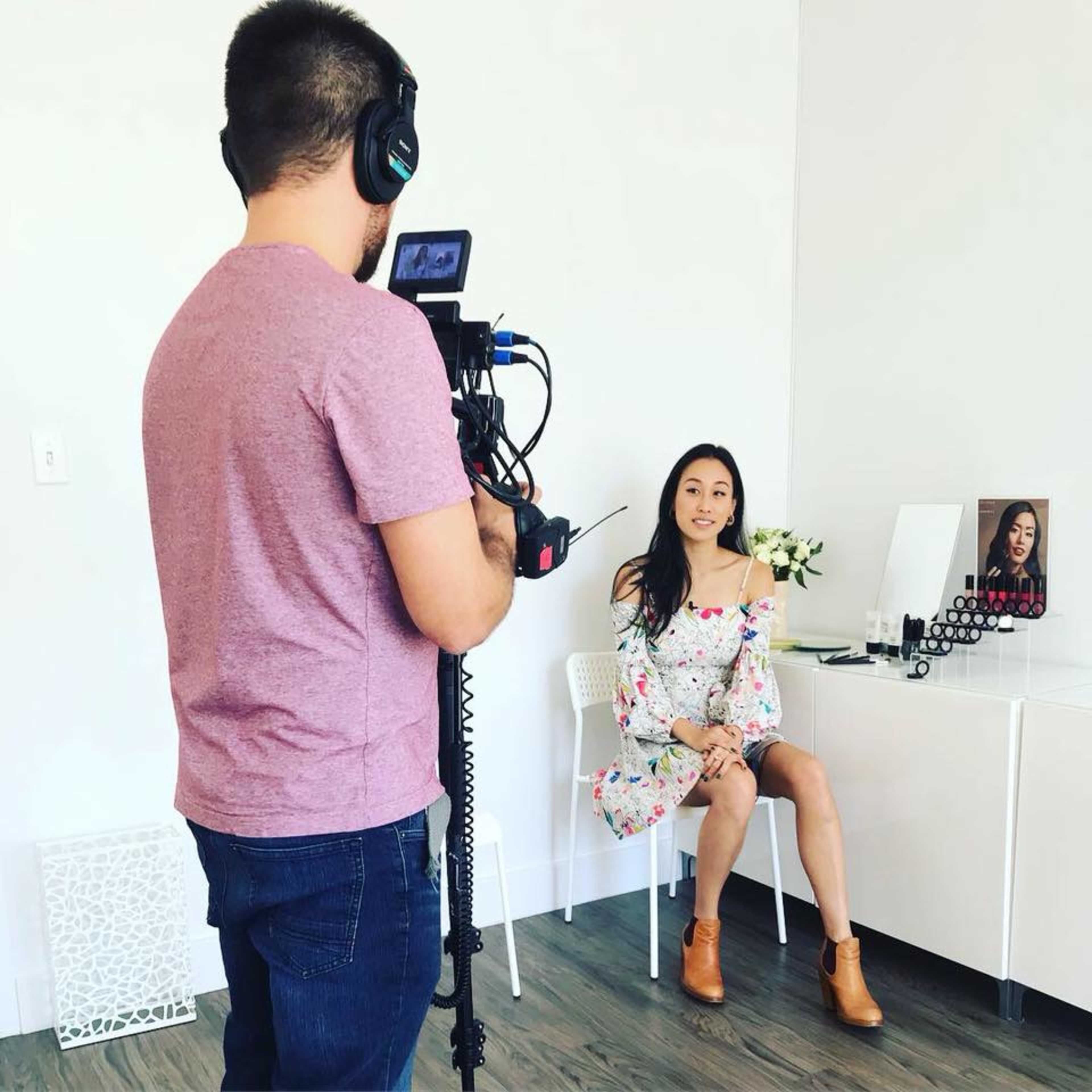 A man films a woman sitting at a table with makeup and a mirror in a bright white room.