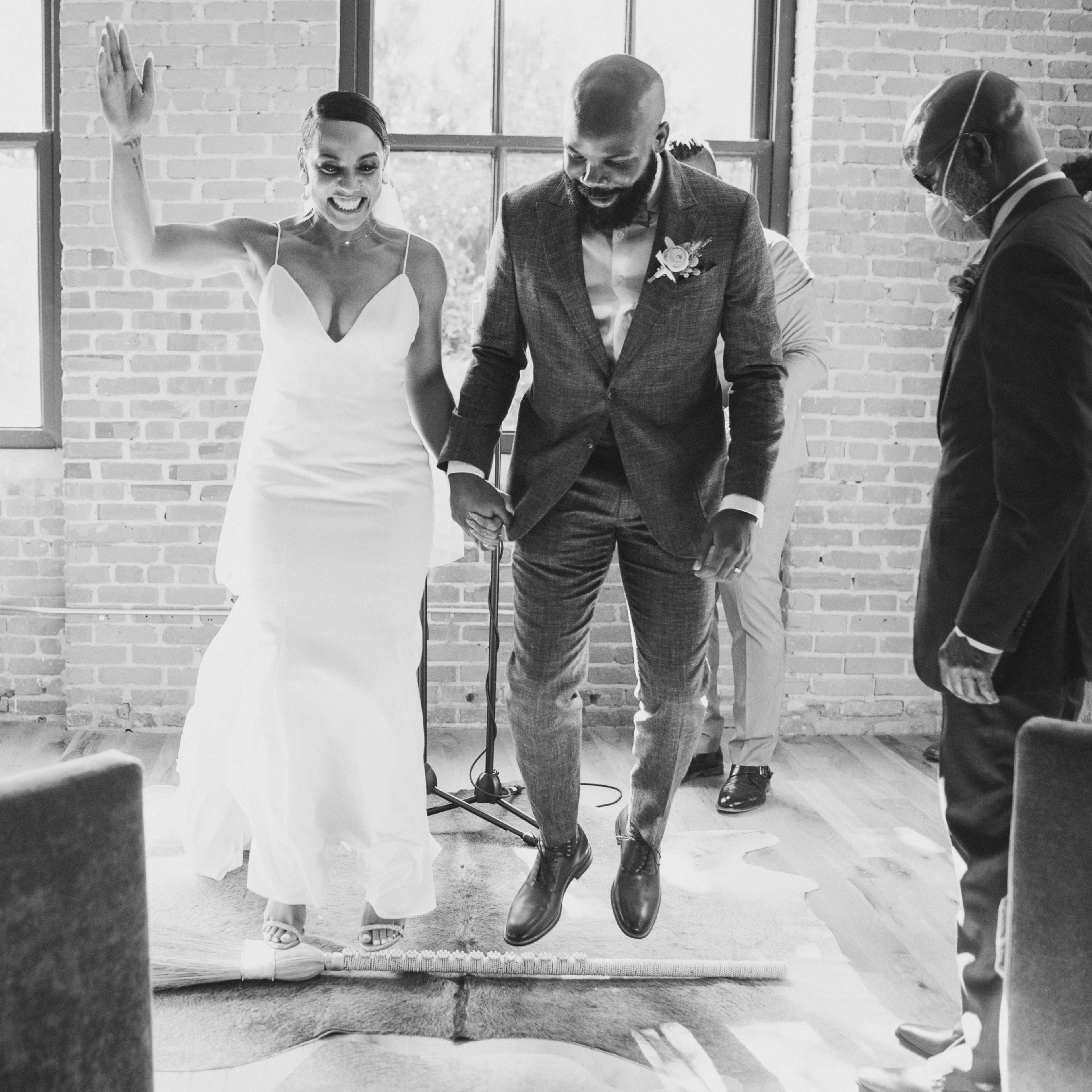 A couple joyfully participates in a wedding ceremony while standing on a rug surrounded by guests in a well-lit room with exposed brick walls.