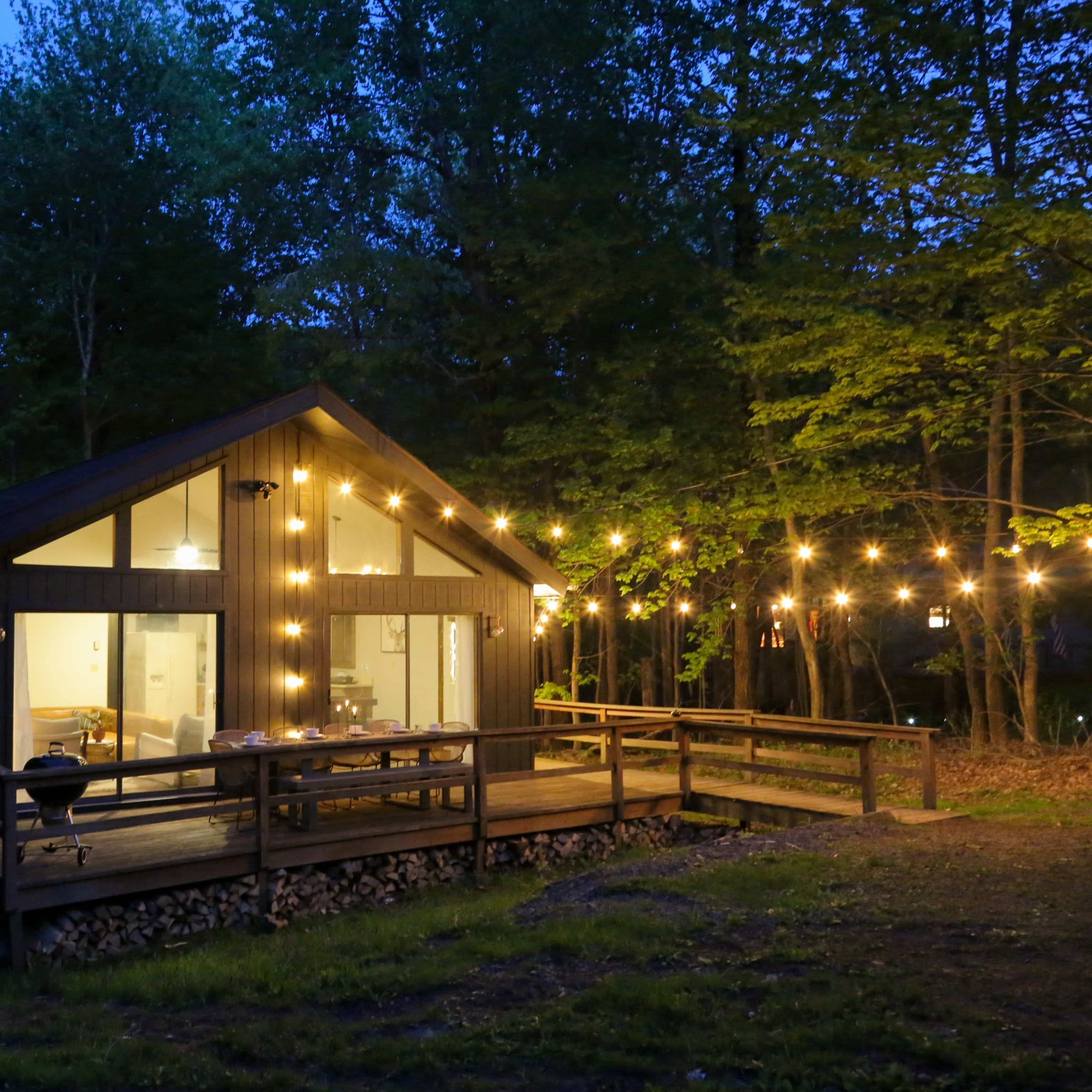 A cabin with large windows and string lights is situated in a wooded area at dusk.