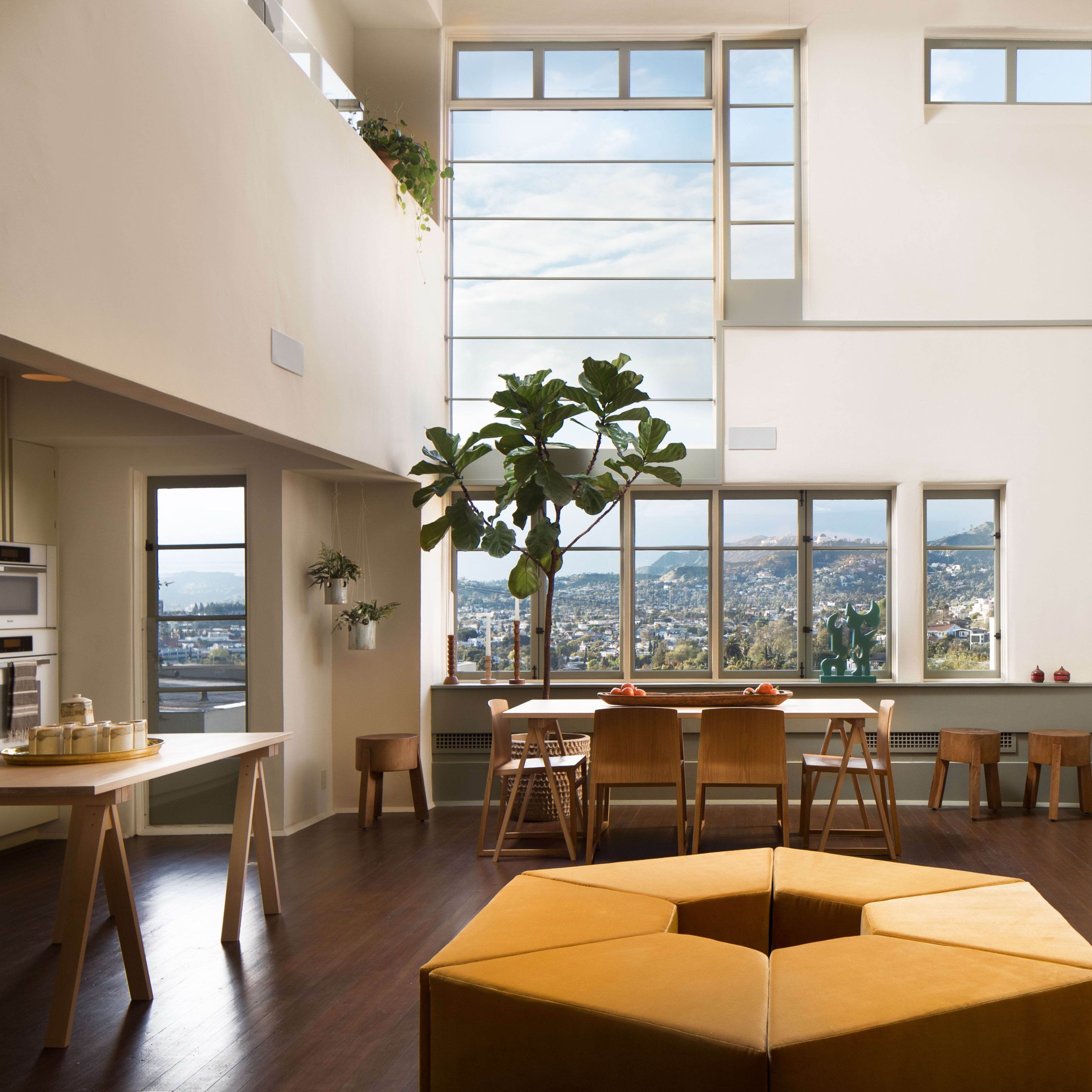A spacious modern living area with large windows, a dining table, and a sectional couch, all featuring minimalist furnishings and a view of the outdoors.