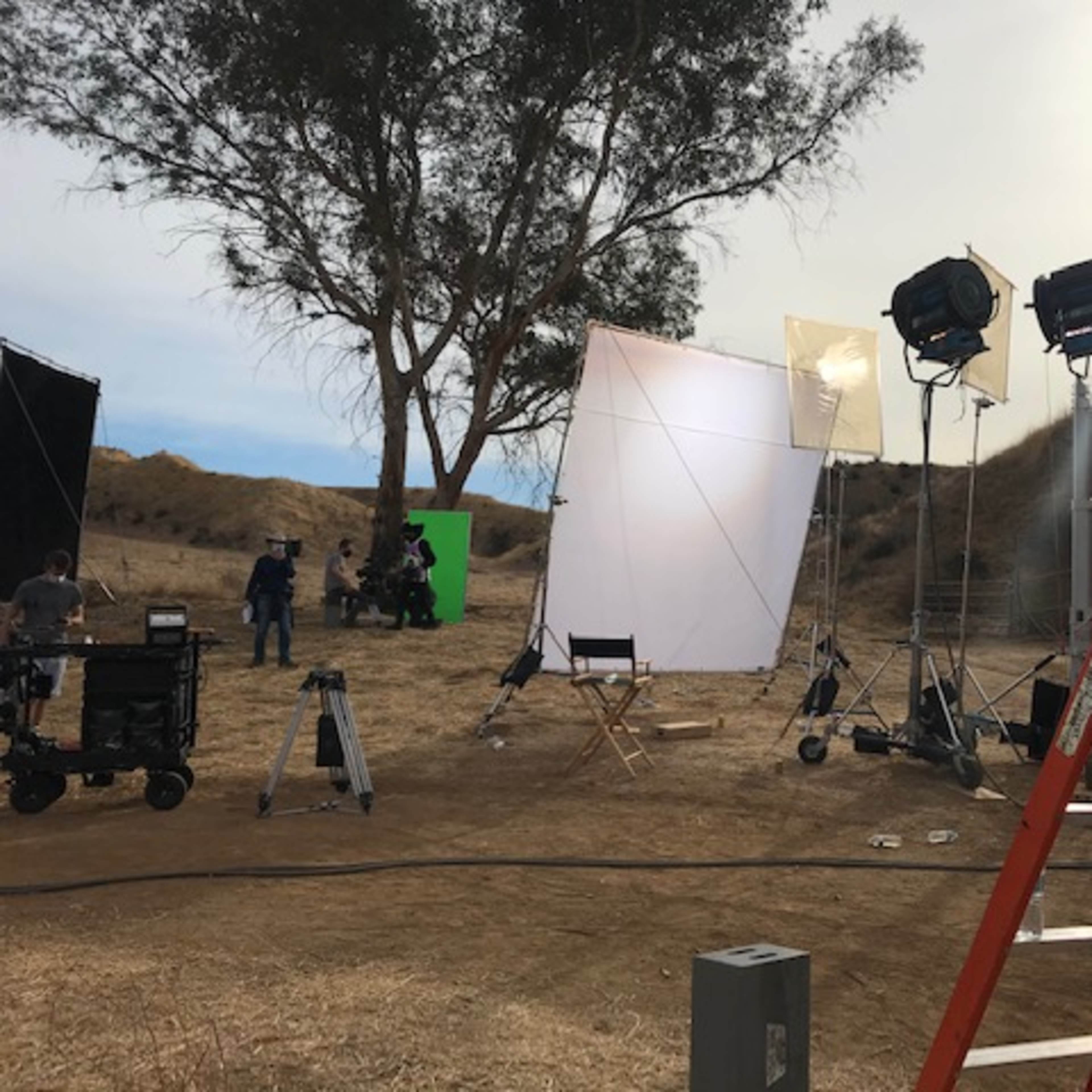 A film set is positioned in a dry outdoor location, featuring lighting equipment, reflectors, and a green screen, with crew members preparing for a shoot.