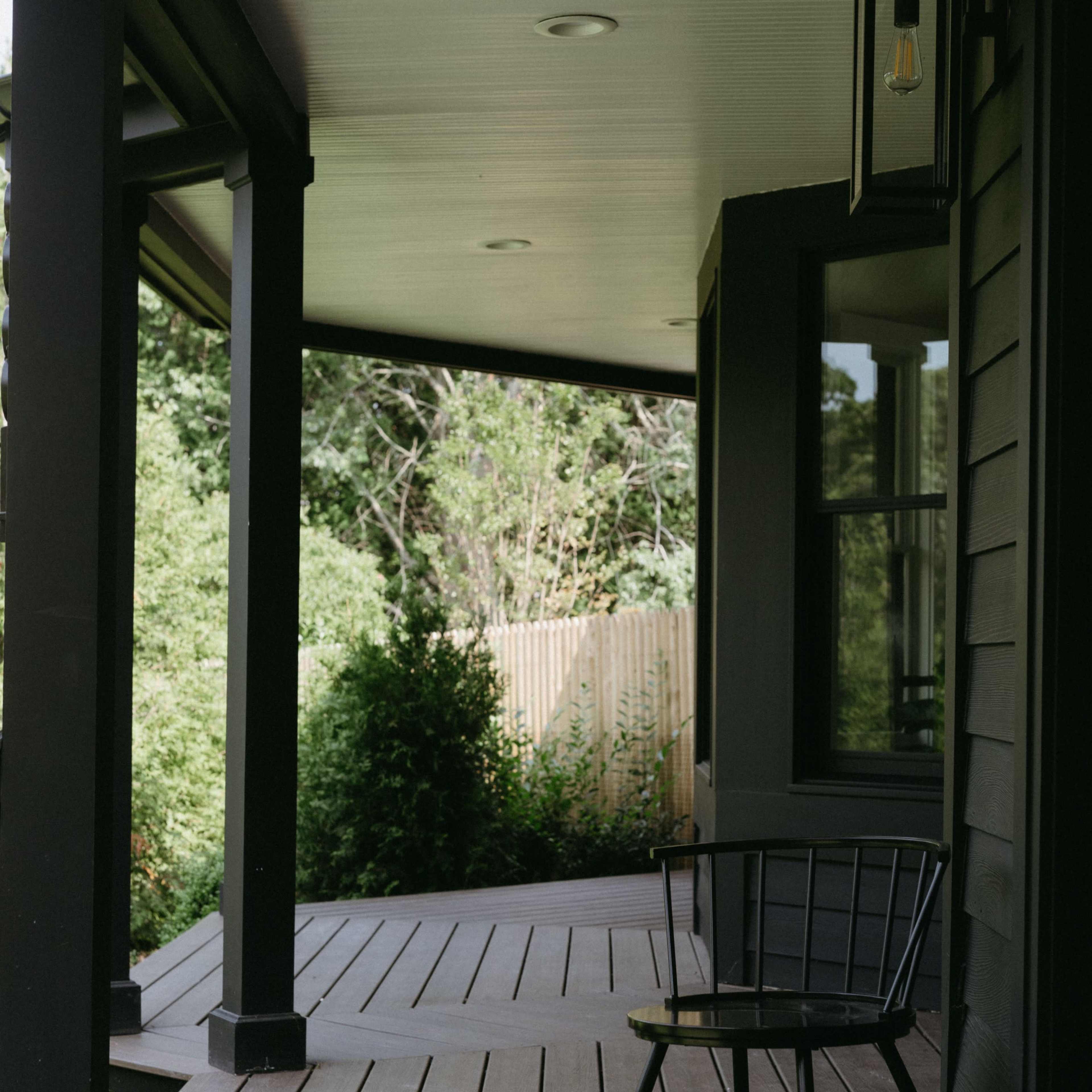 A black chair is positioned on the wooden porch, which features dark beams and a view of greenery through the railing.