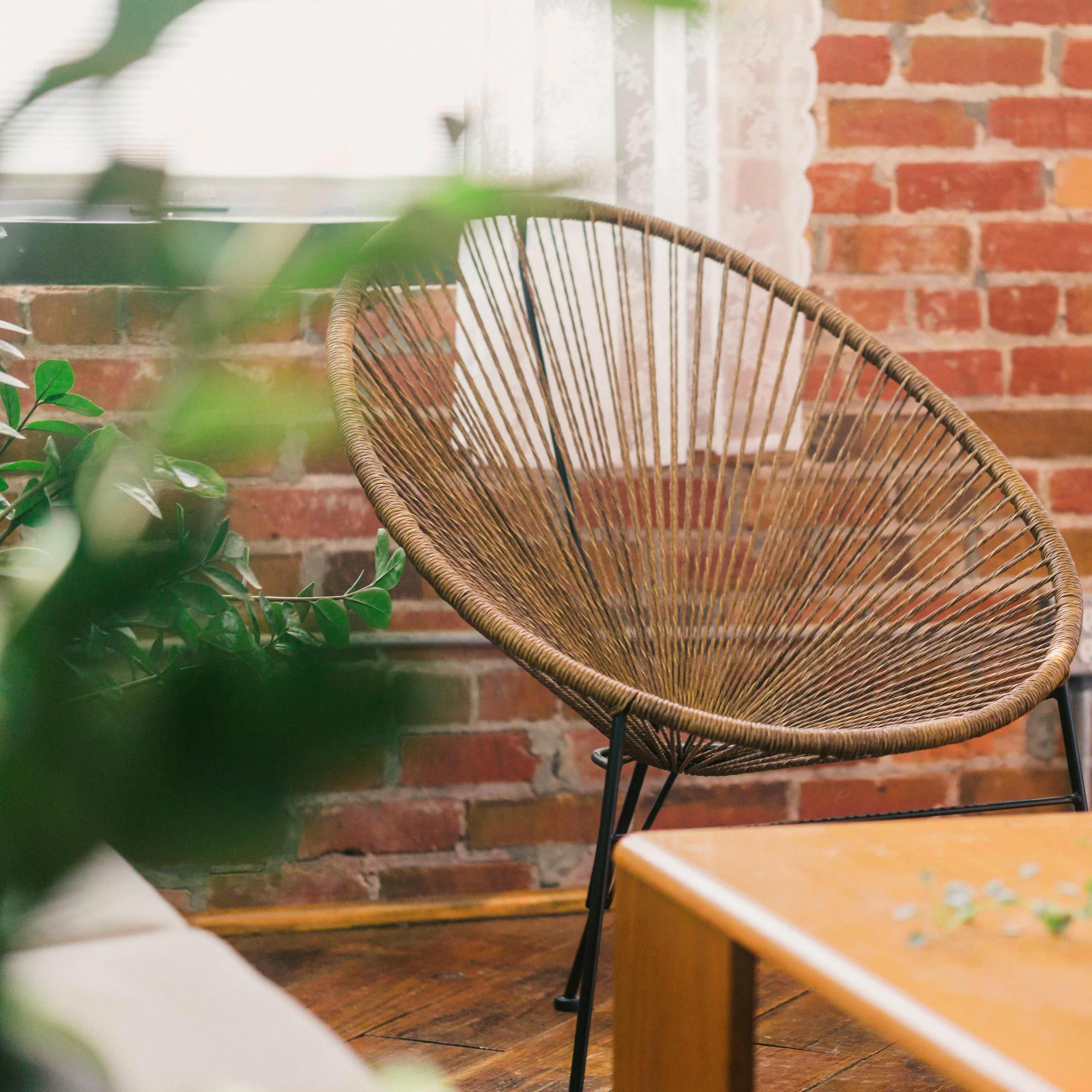 A woven chair is positioned near a wooden table in front of a brick wall, with plants in the foreground.