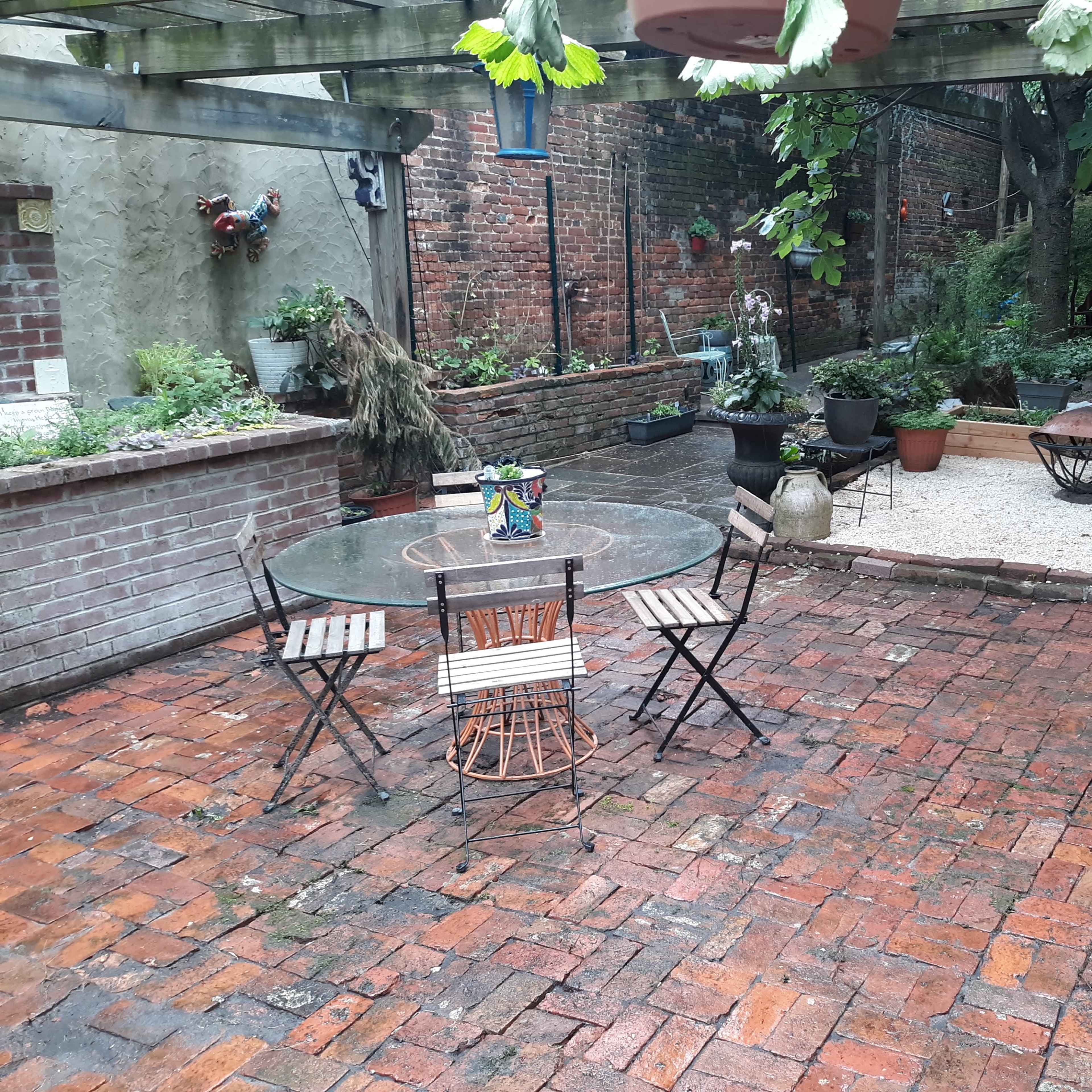 A brick patio with a small round table and chairs, surrounded by potted plants and a gravel area in a garden setting.