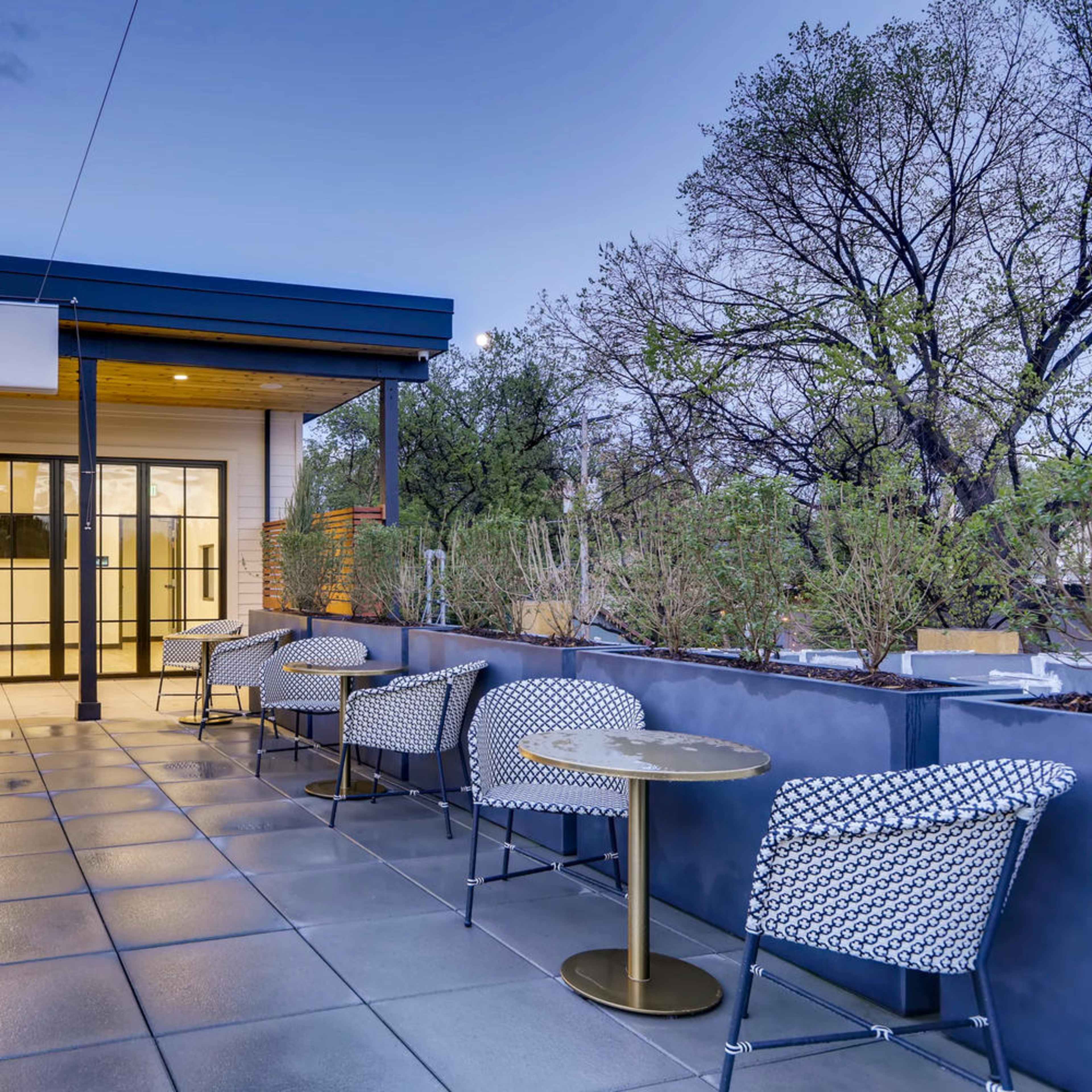 A modern outdoor seating area with multiple tables and chairs, surrounded by greenery and a clear sky at dusk.