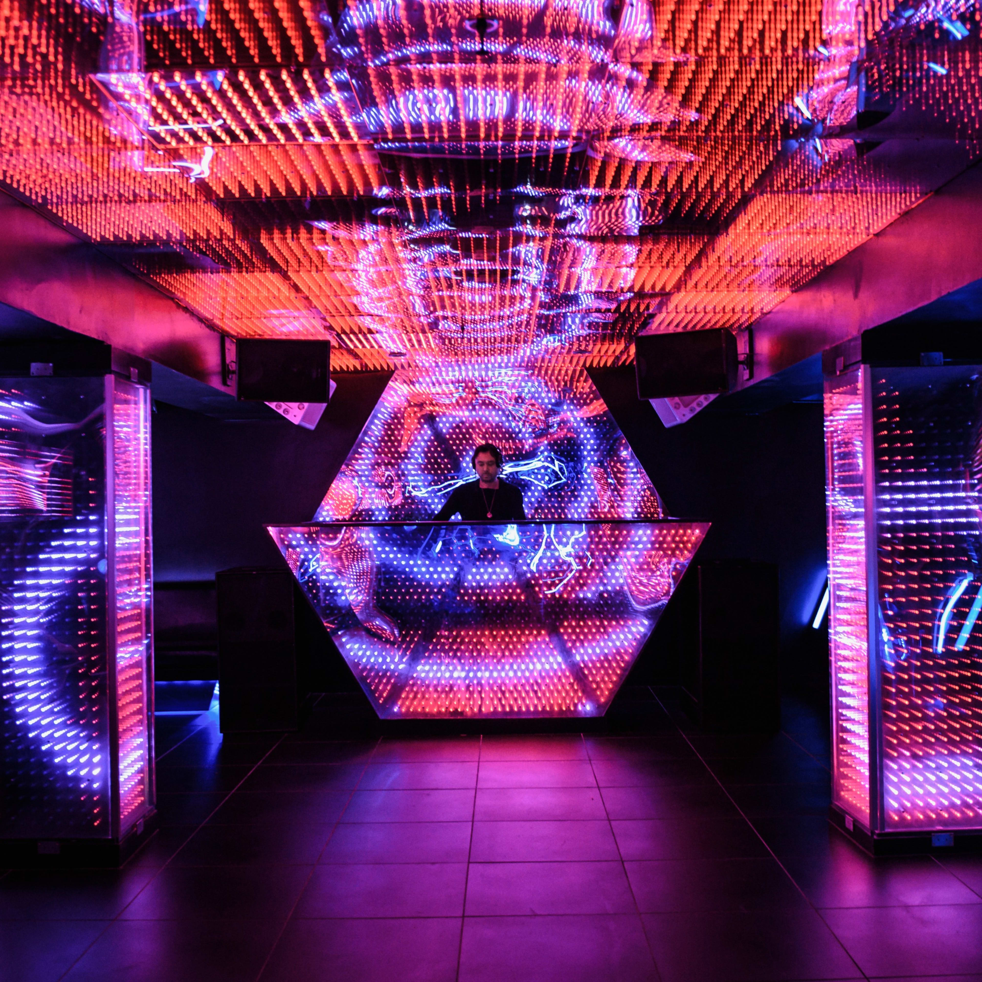 A DJ stands behind a neon-lit hexagonal booth in a vibrant nightclub filled with colorful LED lights.