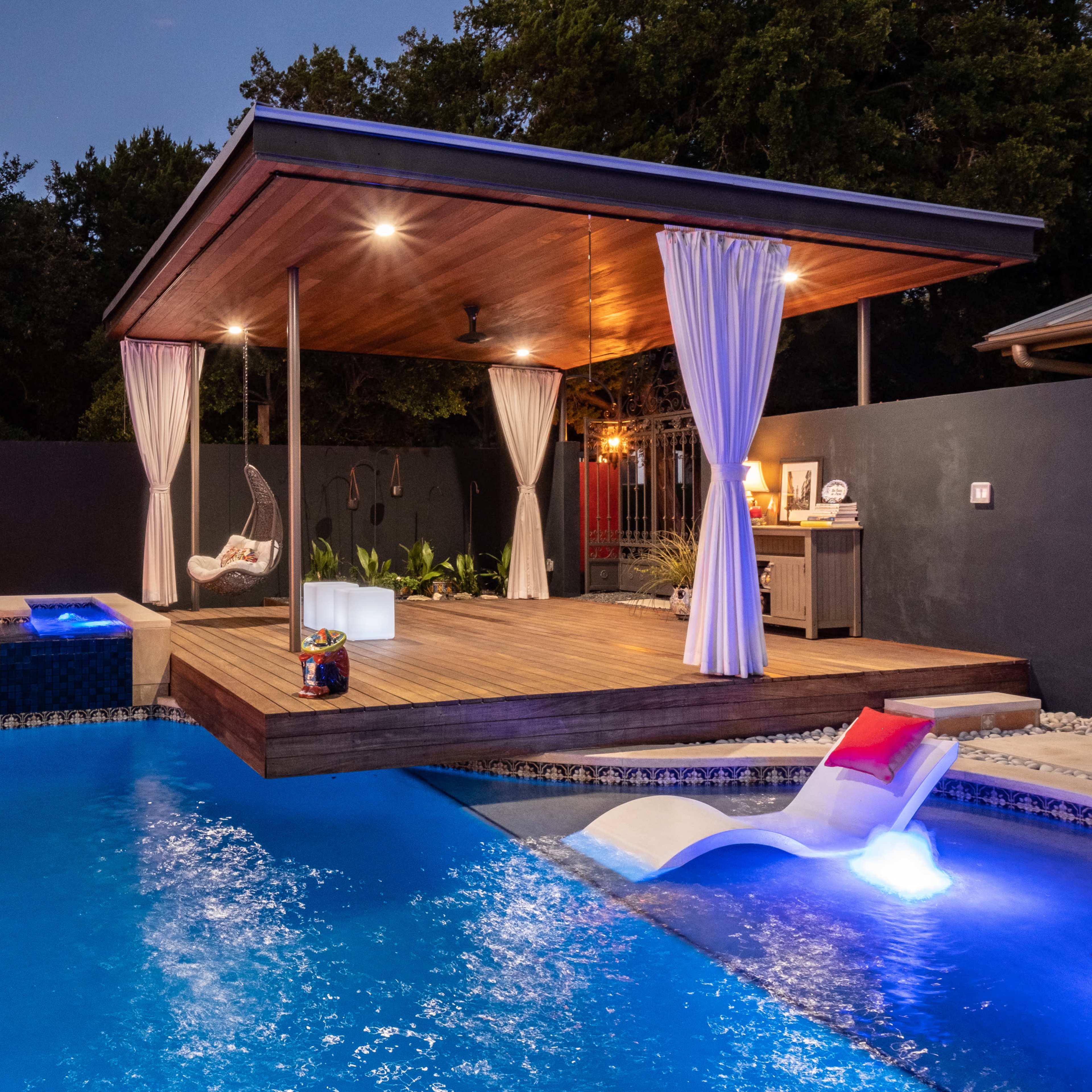 A wooden cabana with draped curtains overlooks a lit pool featuring a spa area, surrounded by trees and a dark wall with vertical lights.