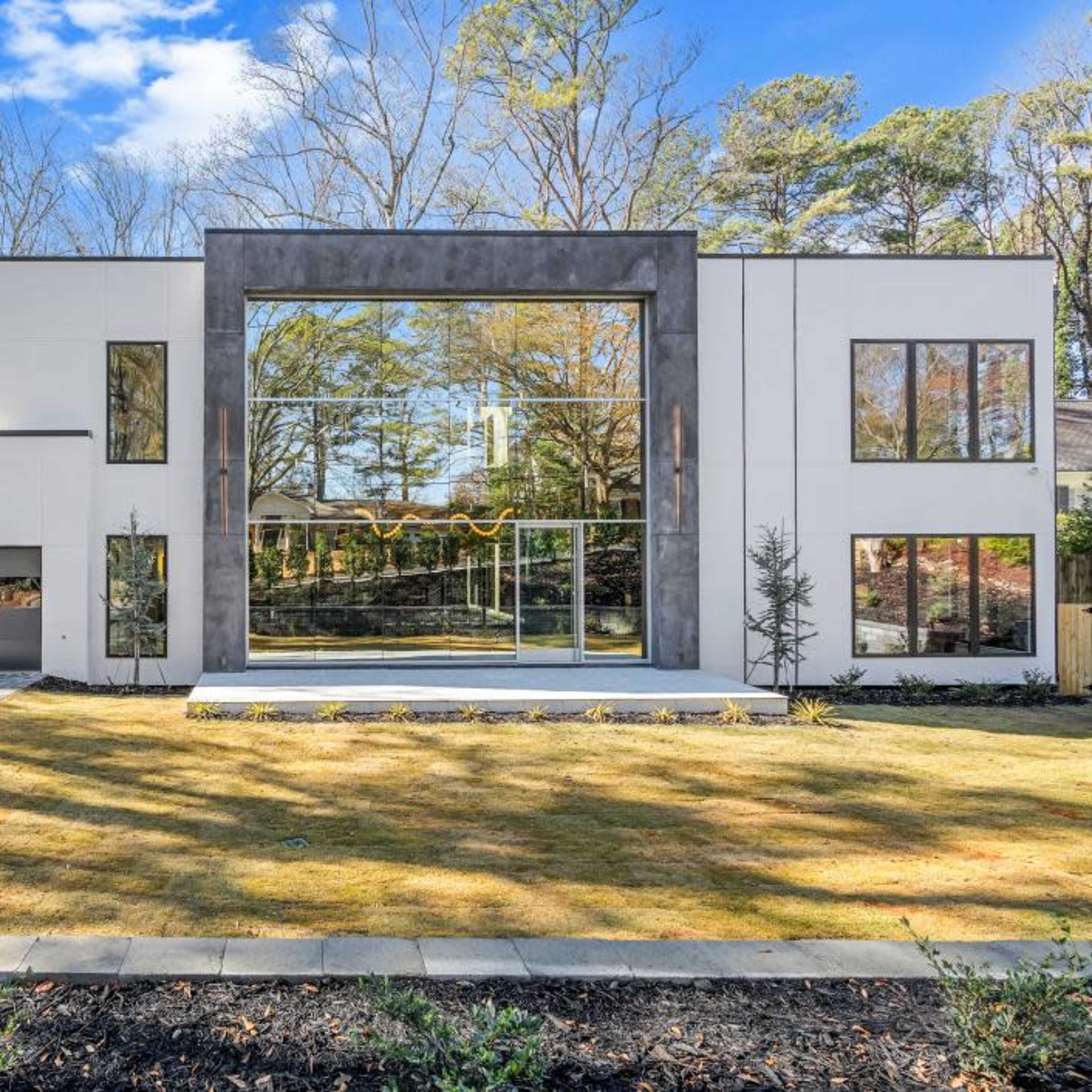 A modern two-story house with large glass windows and a landscaped front yard is set against a backdrop of trees.