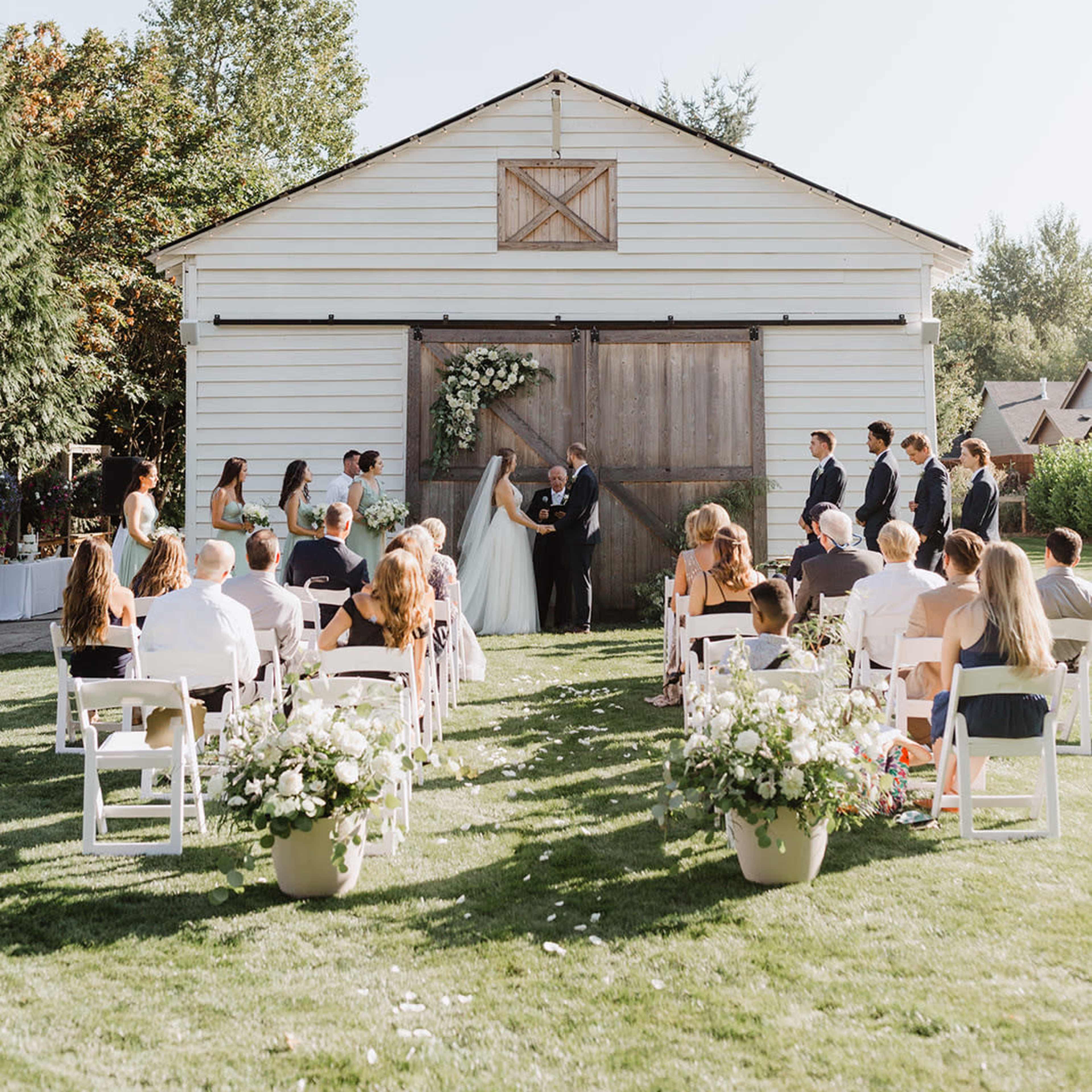 A couple stands under a floral arch in front of a large barn while guests seated on white chairs observe the wedding ceremony.