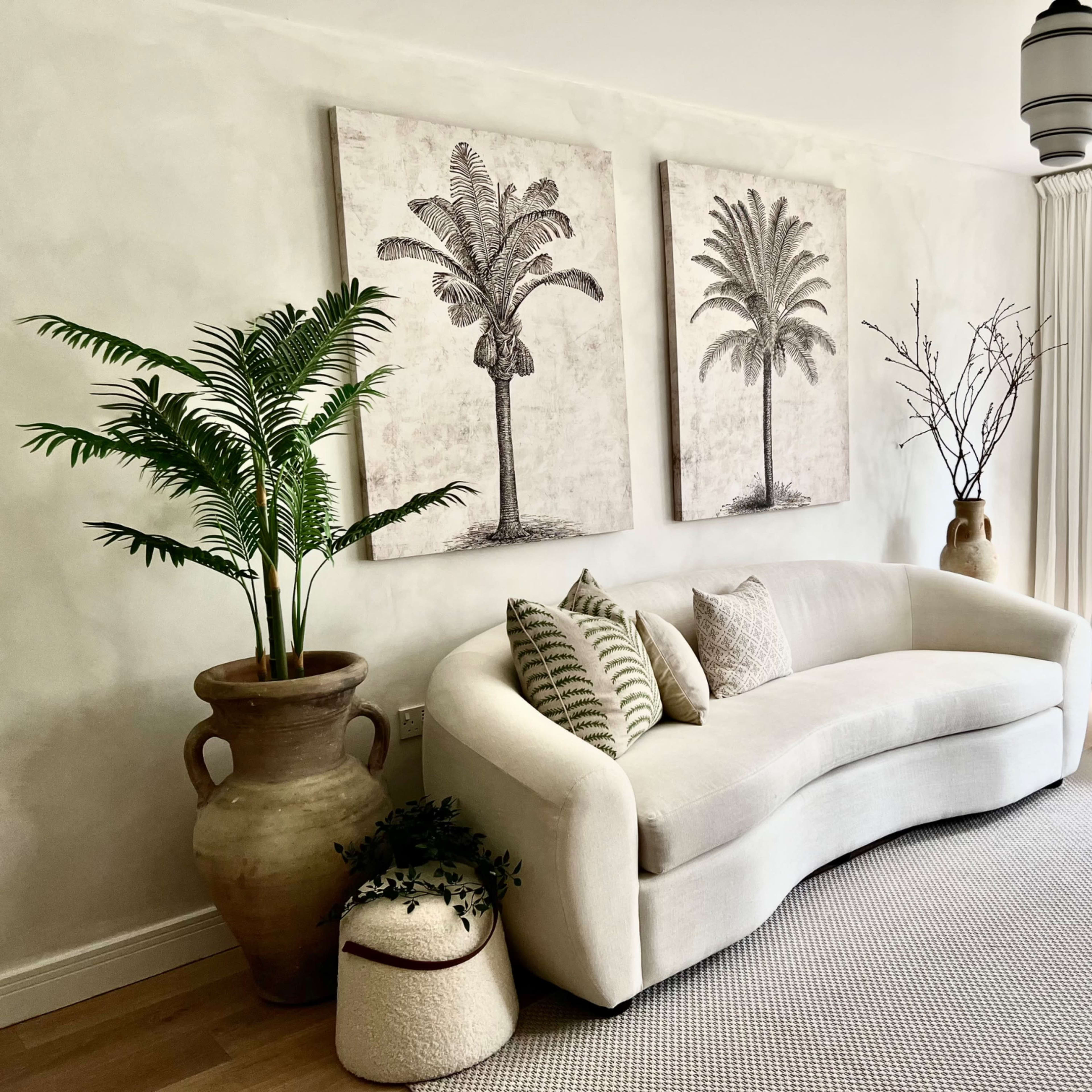 A light-colored living room features a curved sofa, decorative palm prints on the wall, and various plants in stylish pots.