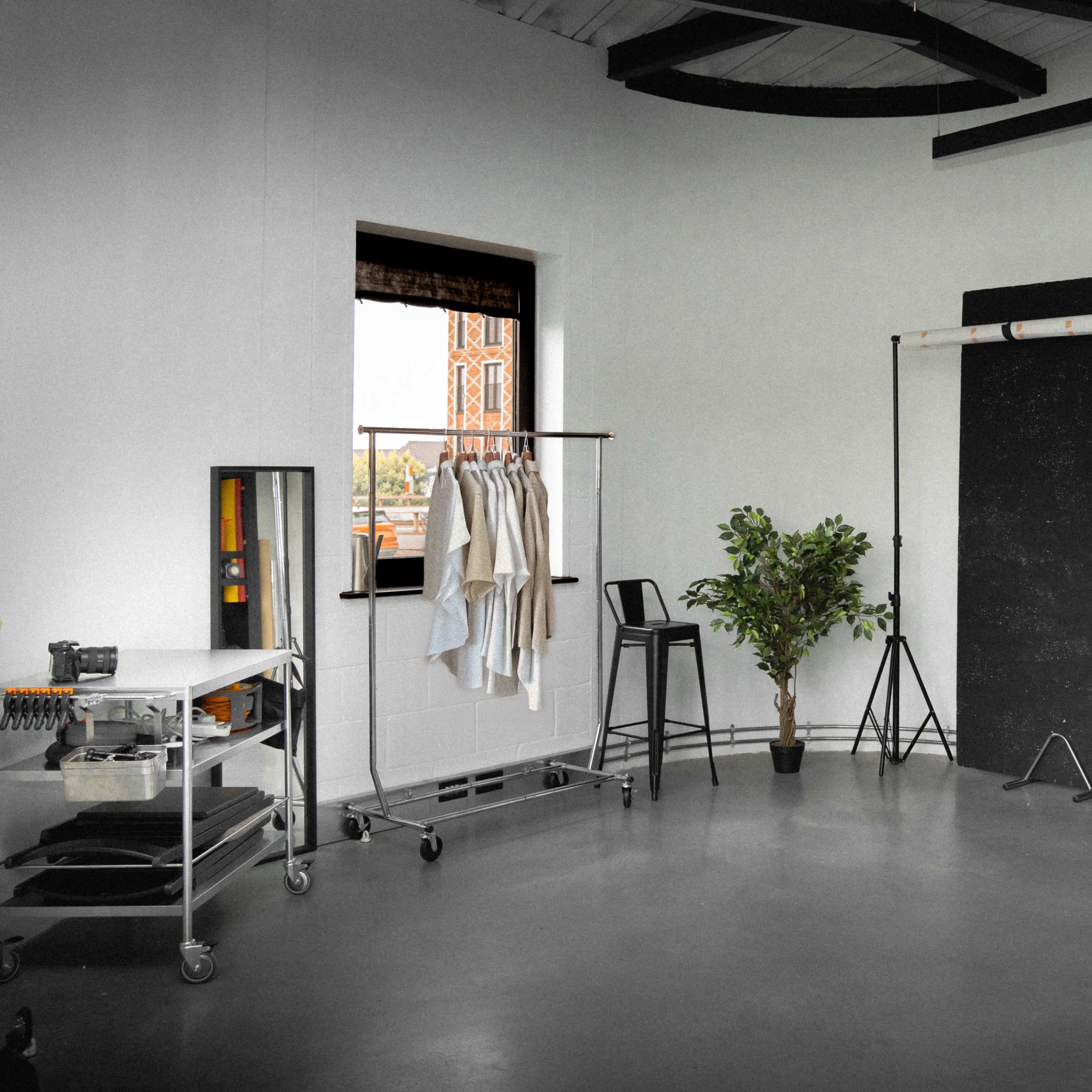 A minimalist studio space features a window, black and white backdrops, a clothing rack, a potted plant, and photography equipment.