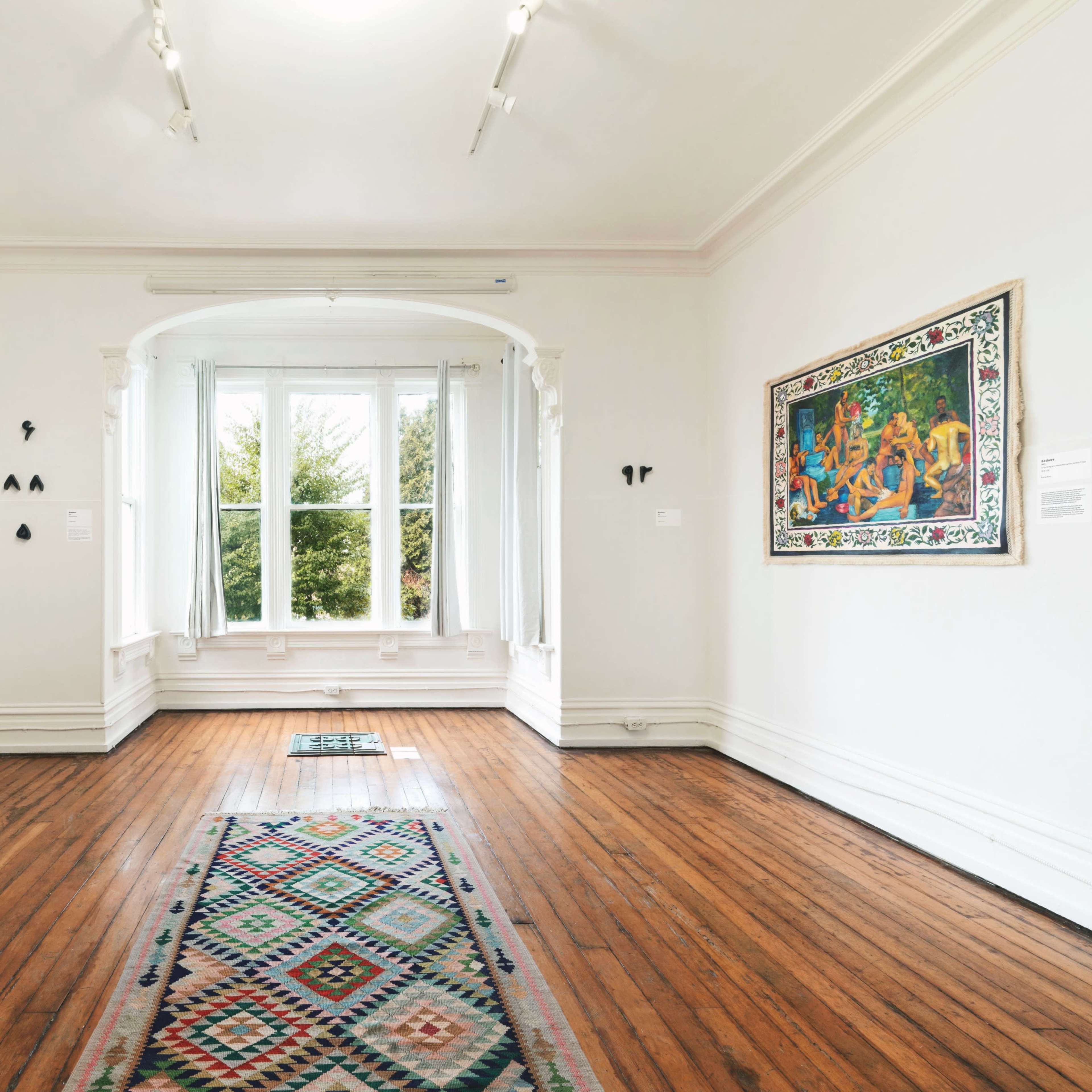 A spacious art gallery features hardwood floors, a central rug, and several paintings displayed on the white walls, with large windows allowing natural light to fill the room.