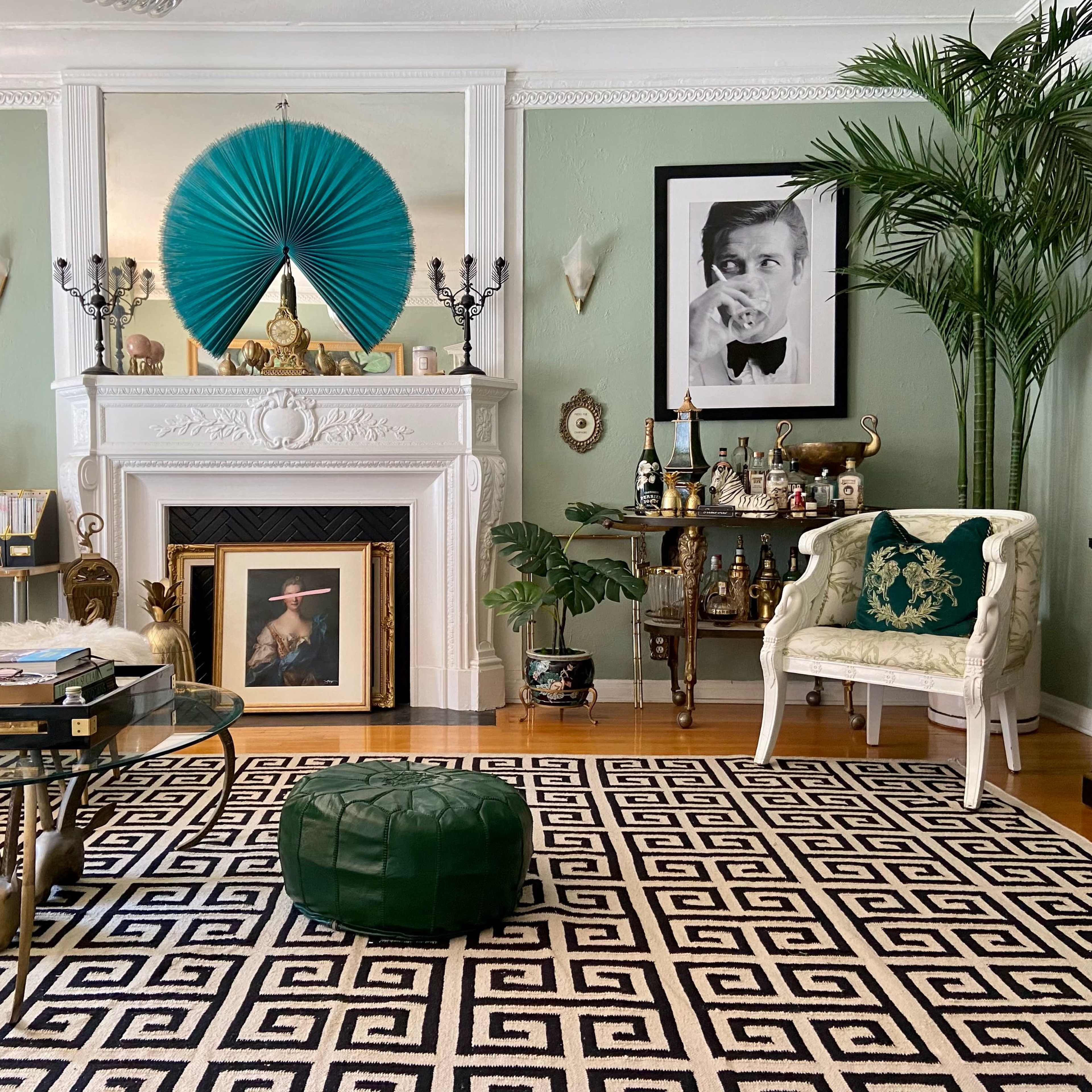 The room features a stylish living area with a black and white geometric rug, a white fireplace adorned with decorative items, and a green velvet ottoman.