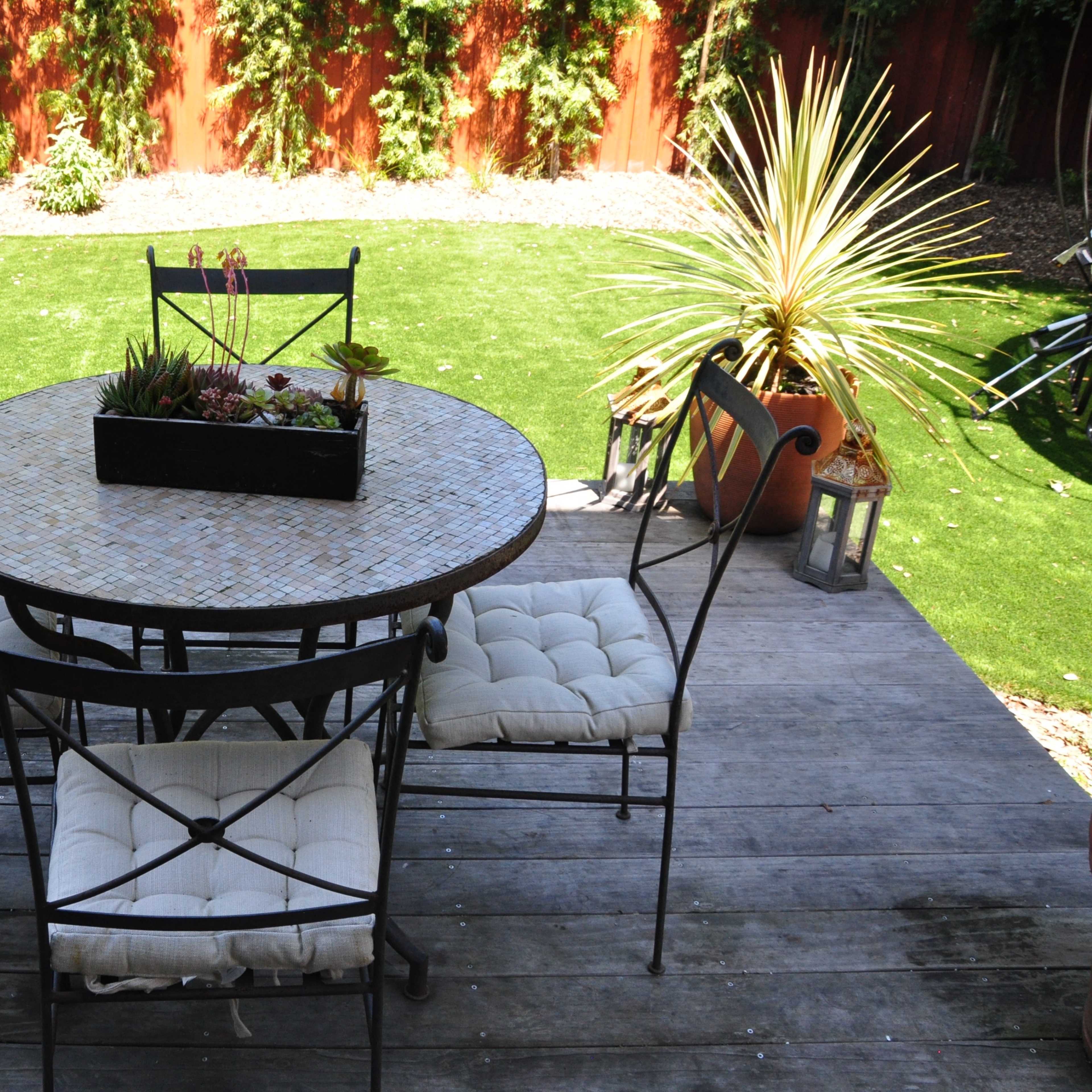 A round table with four chairs is set on a wooden deck surrounded by greenery and decorative plants.
