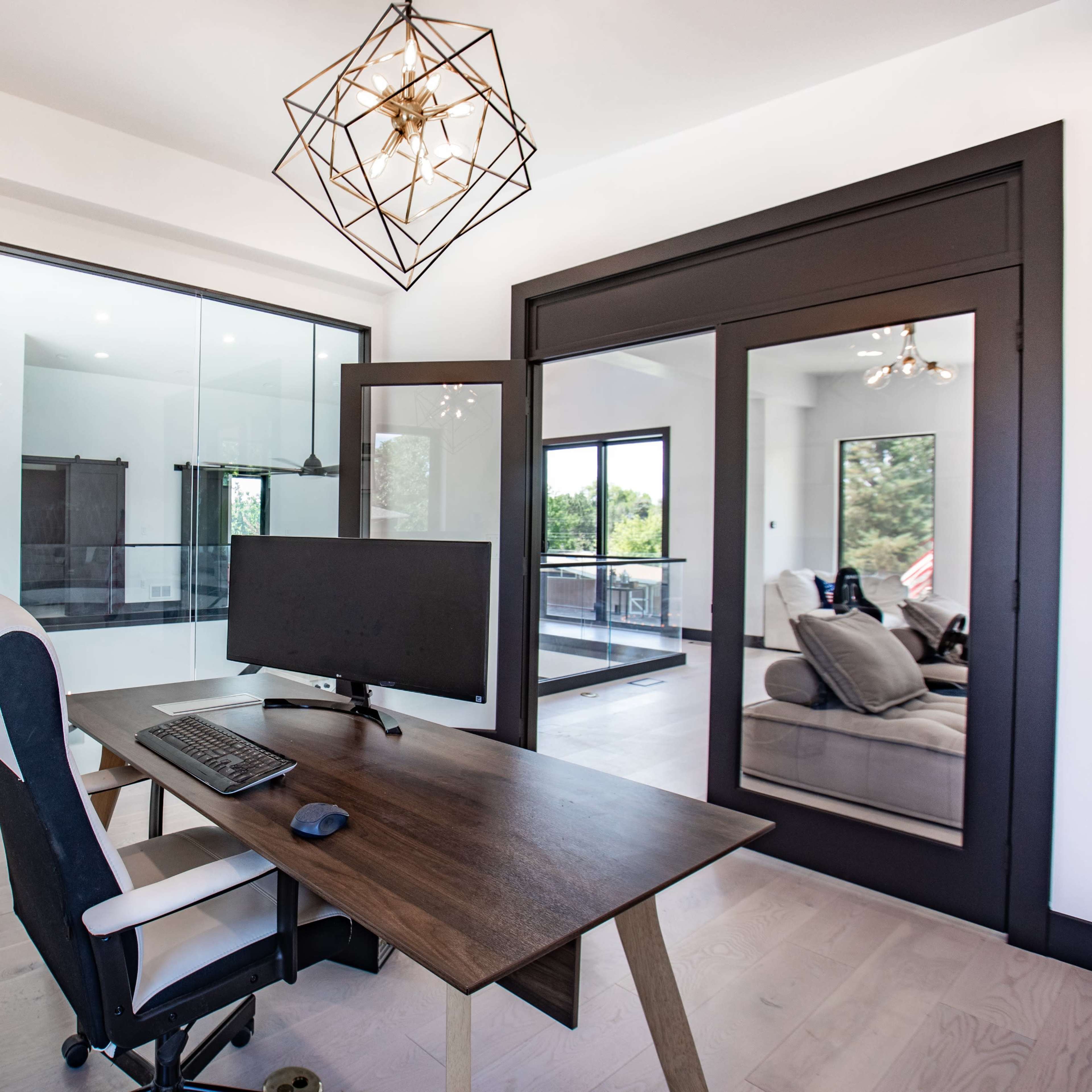 A modern home office features a wooden desk with a monitor and an ergonomic chair, enclosed by glass walls showcasing a bright interior.