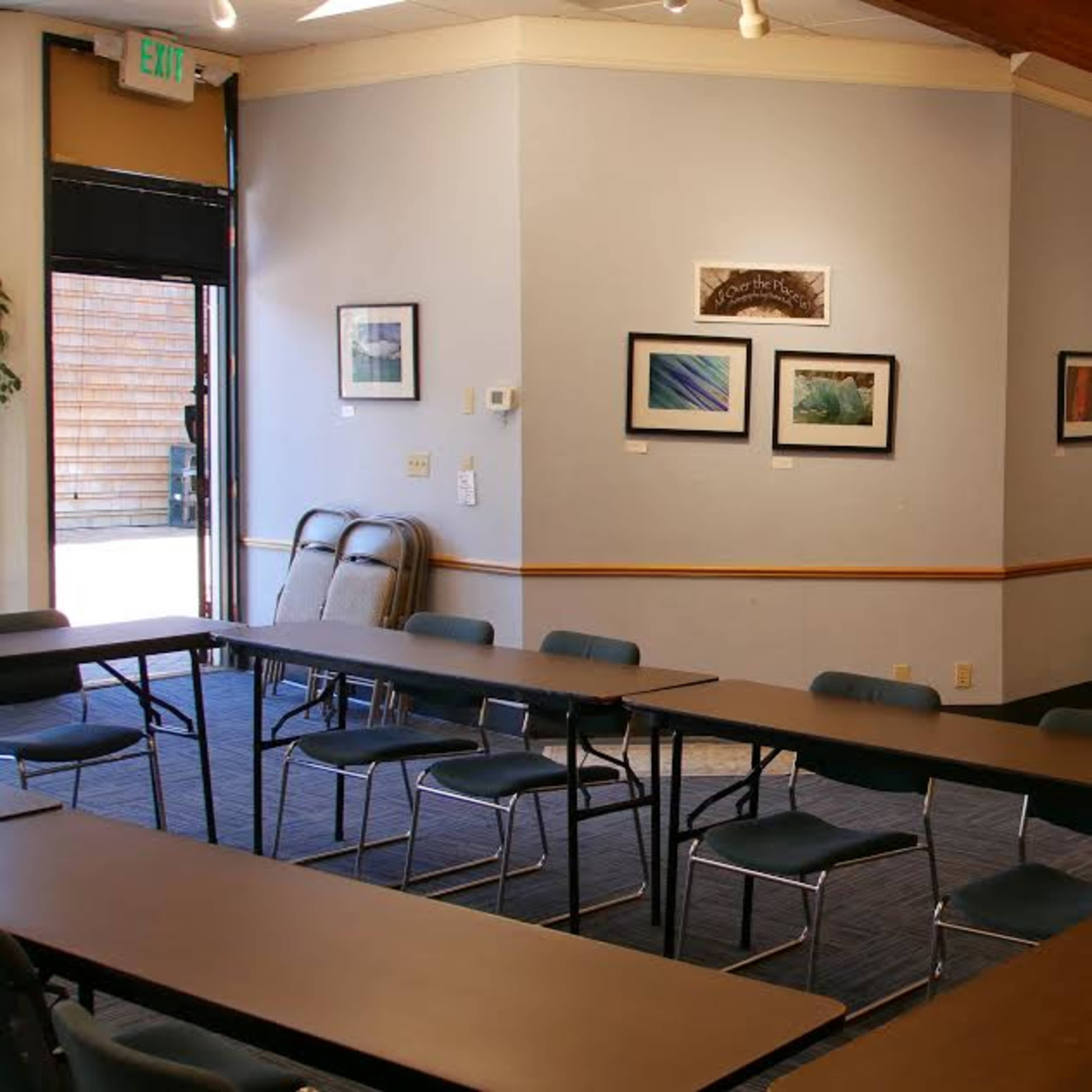 A small meeting room with two long tables arranged in an L-shape, surrounded by chairs, and featuring framed artwork on the walls.