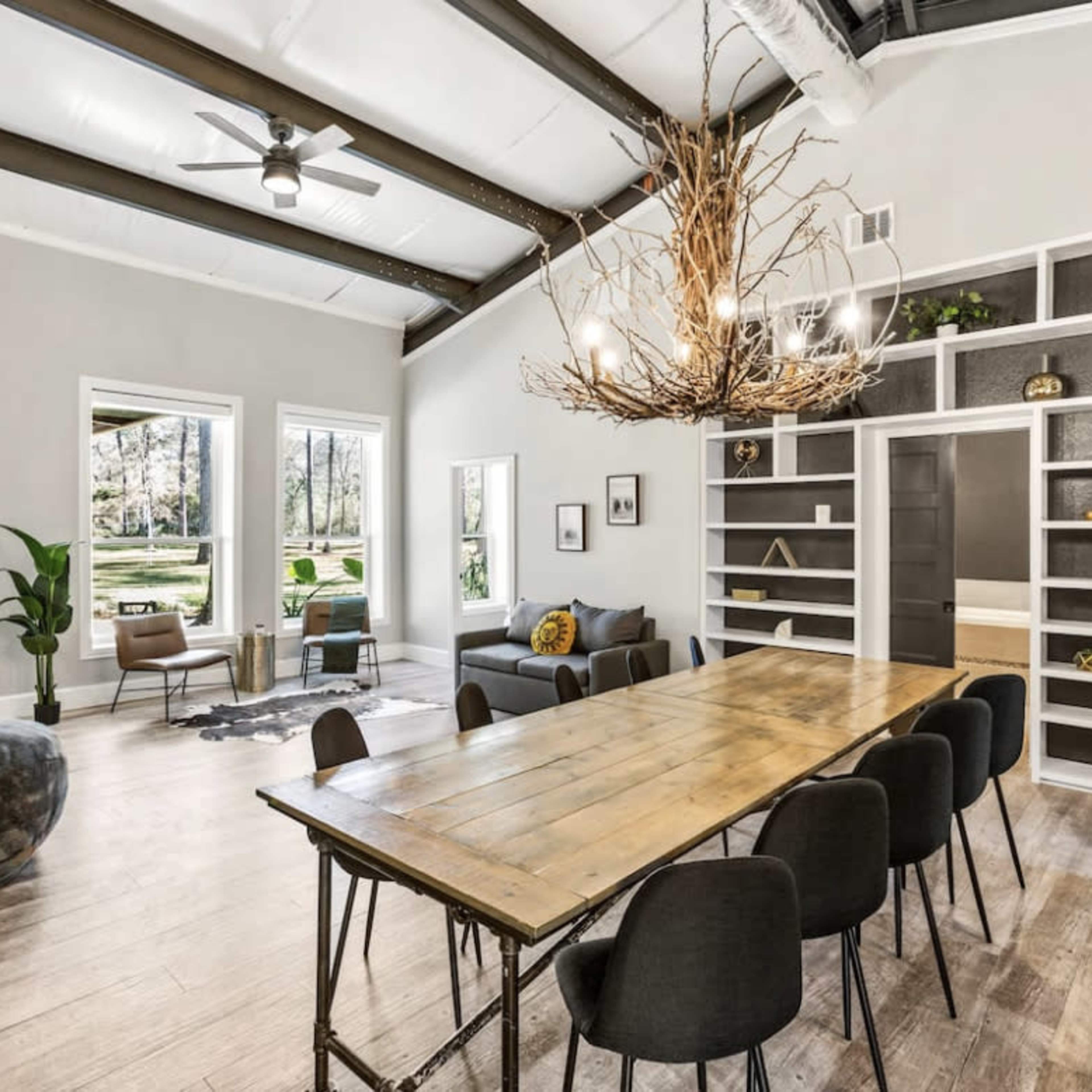 A spacious living area features a large wooden dining table, black chairs, and a comfortable seating area with large windows letting in natural light.