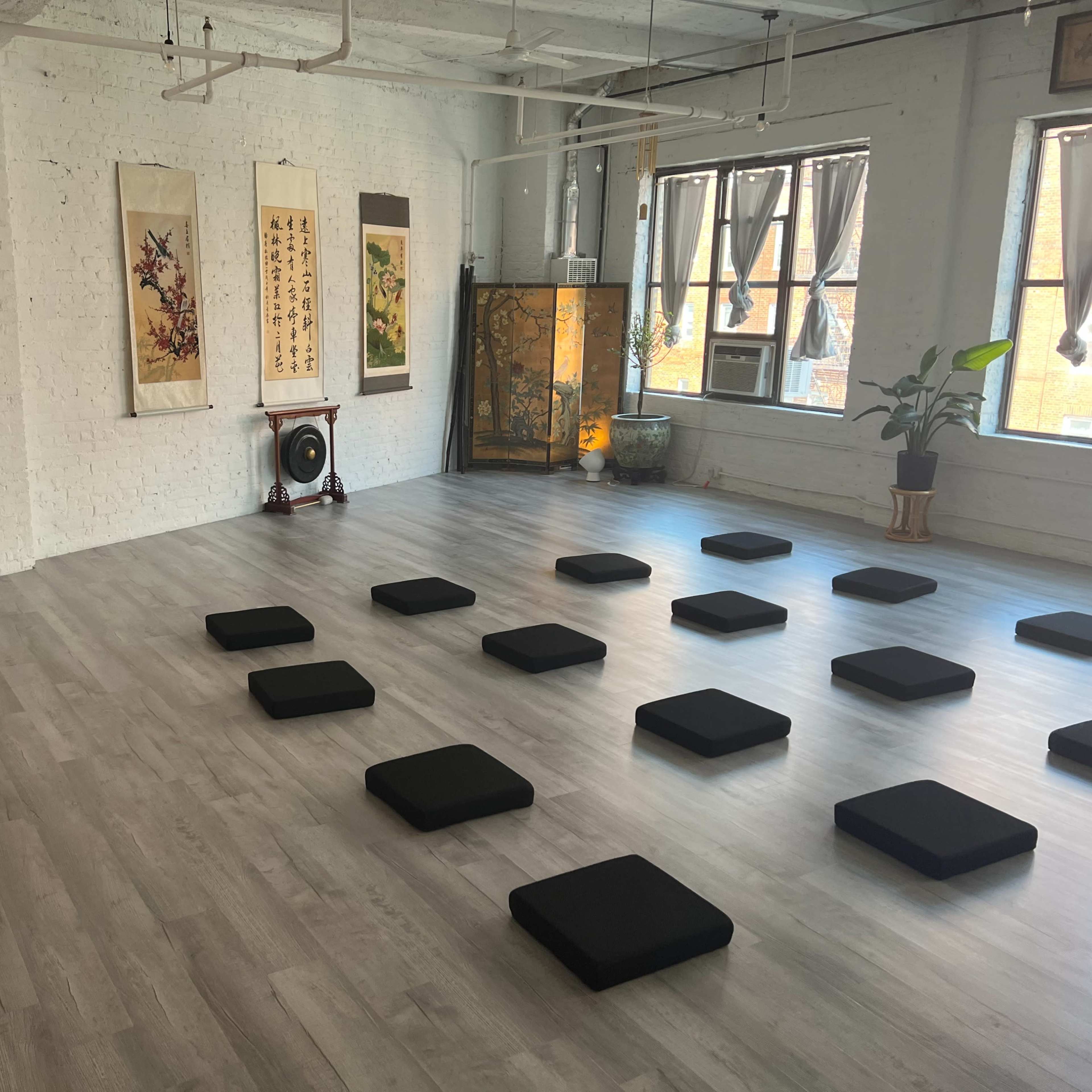 A spacious room set up for meditation, featuring several black cushions arranged on a light wooden floor, with decorative paintings and a plant in the background.