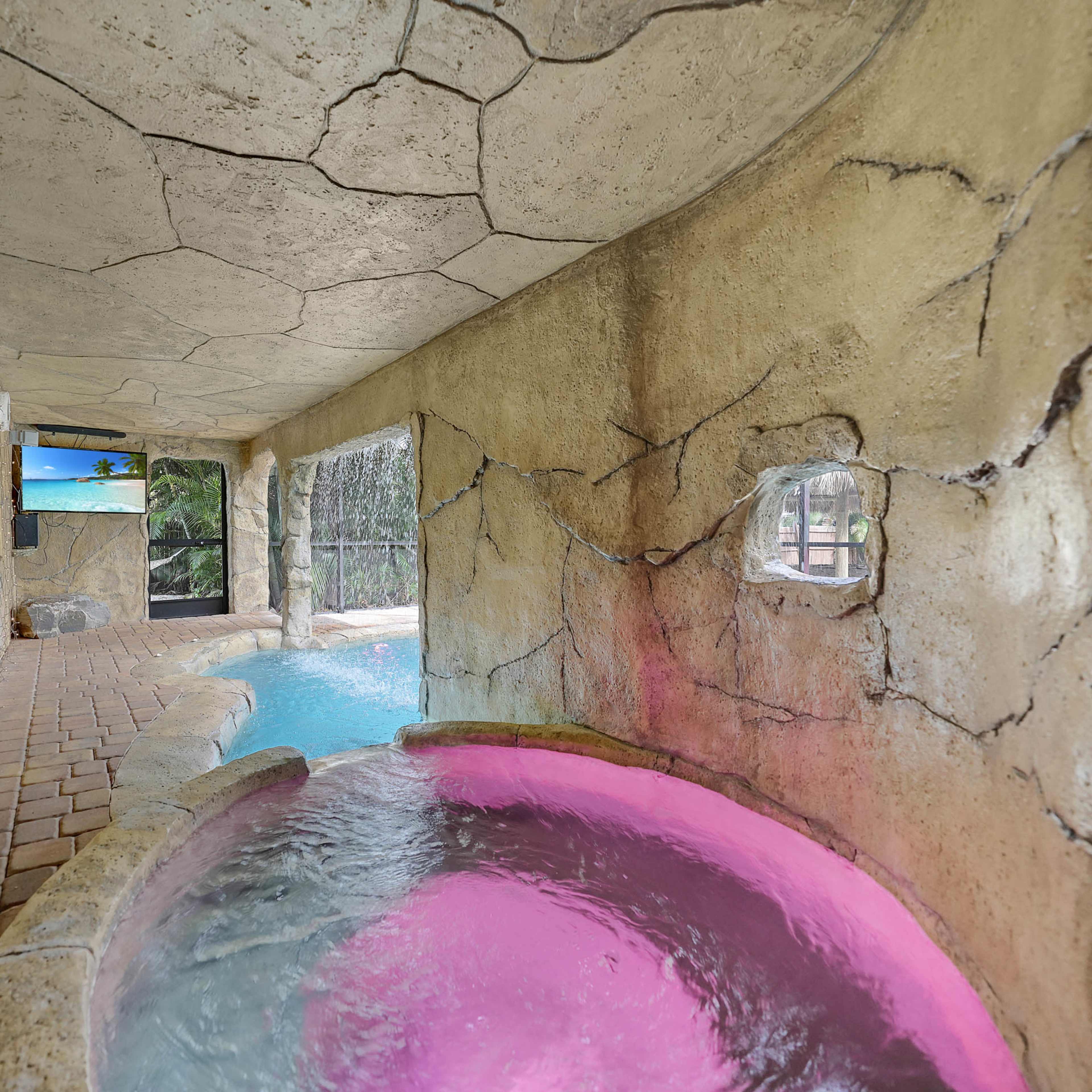 A stone-walled indoor space featuring a small hot tub with pink lighting adjacent to a swimming pool area.