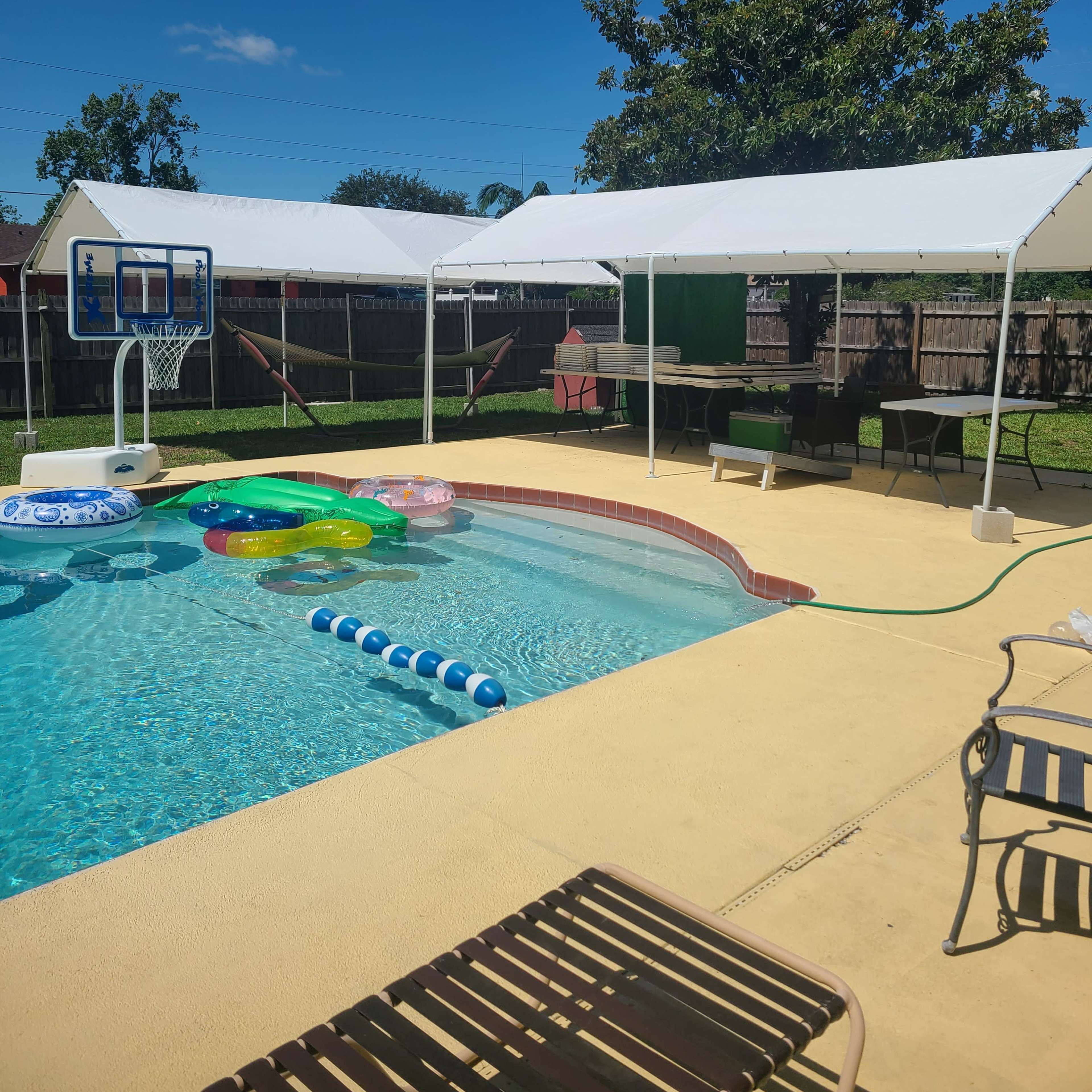 A backyard pool area with a basketball hoop, several floating toys in the water, and two large tents set up beside the pool.
