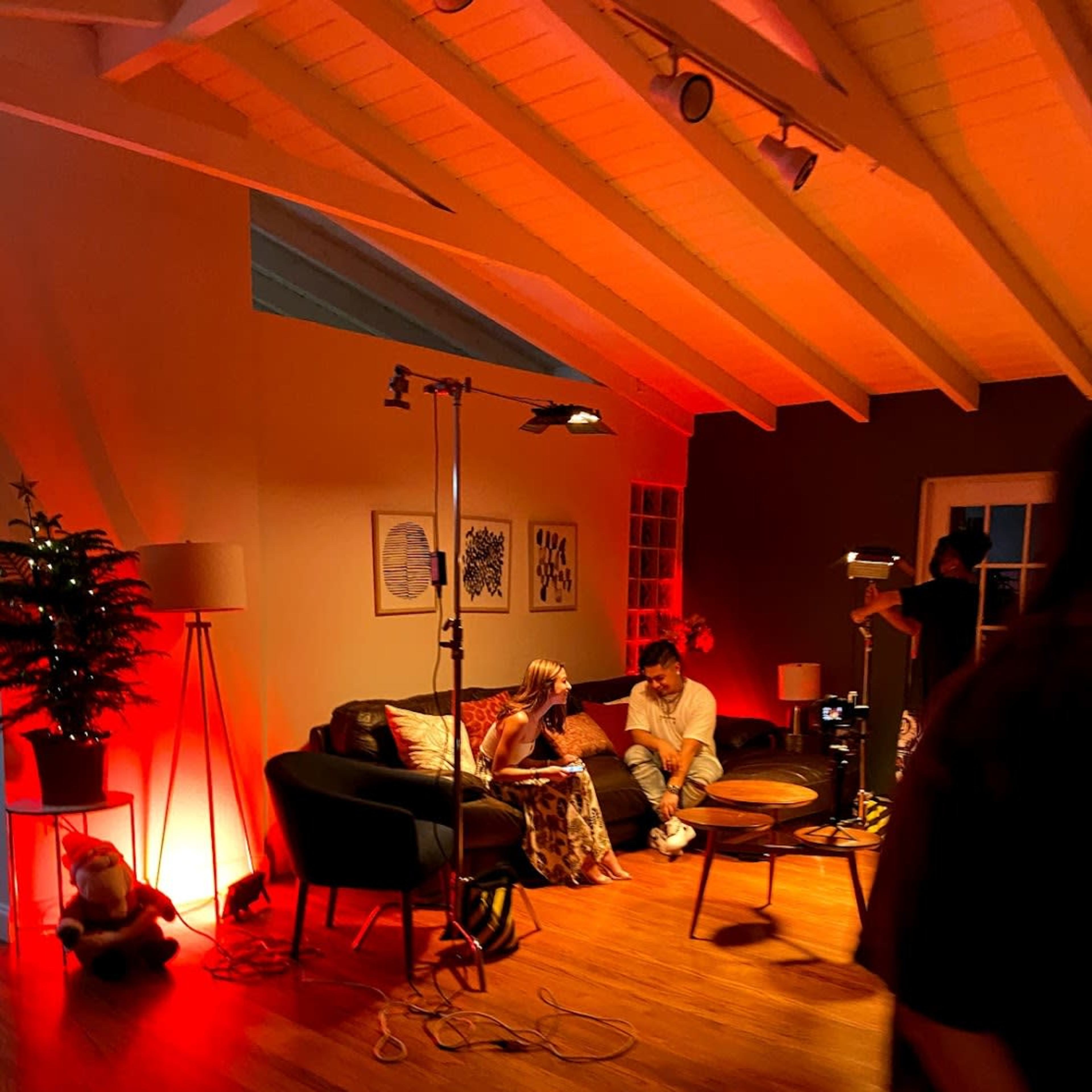 The scene depicts a cozy room with two individuals seated on a couch under warm red lighting, surrounded by various lighting equipment and a decorative tree.