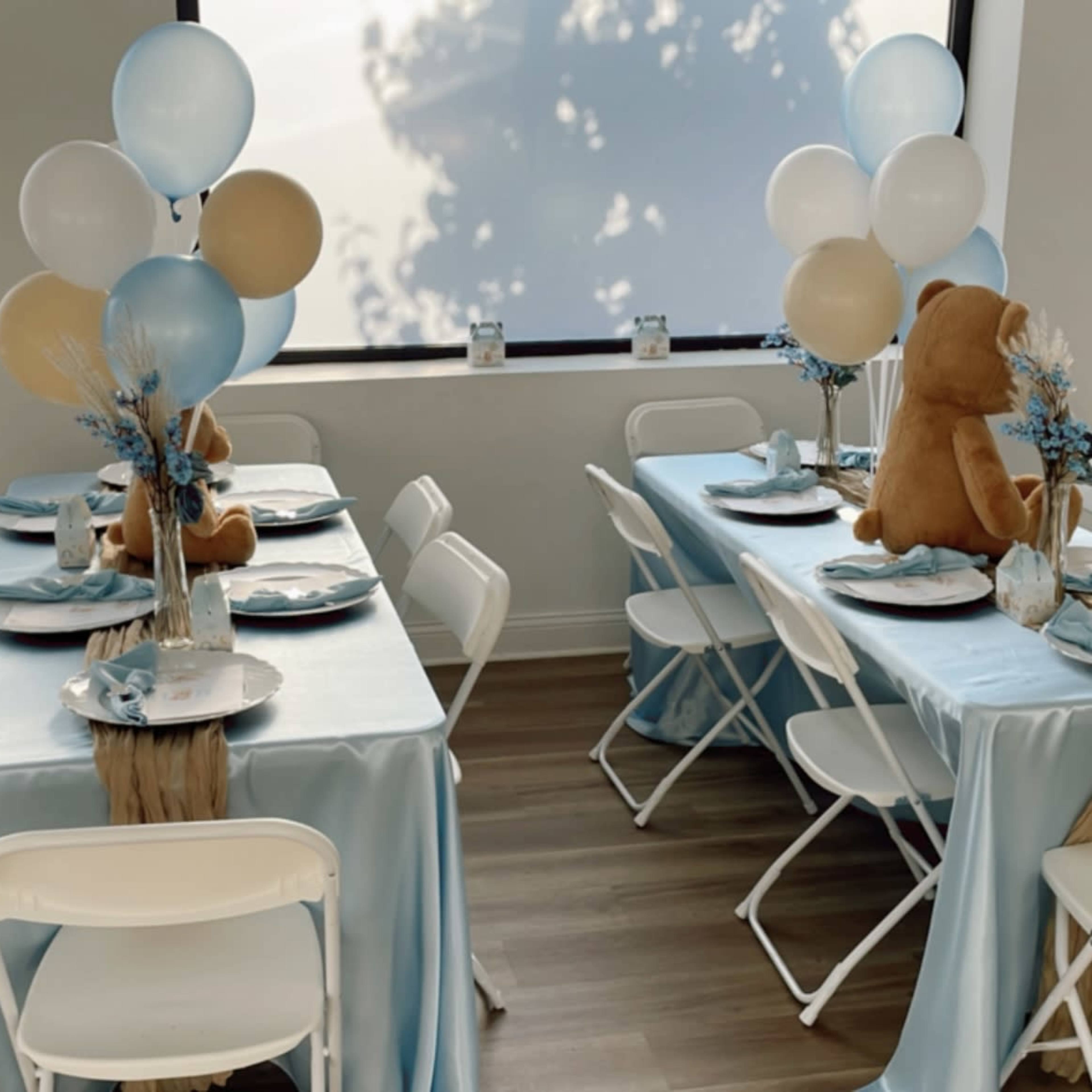 A decorated event space with light blue tablecloths, white folding chairs, and tables set for an occasion featuring balloons and a bear centerpiece.