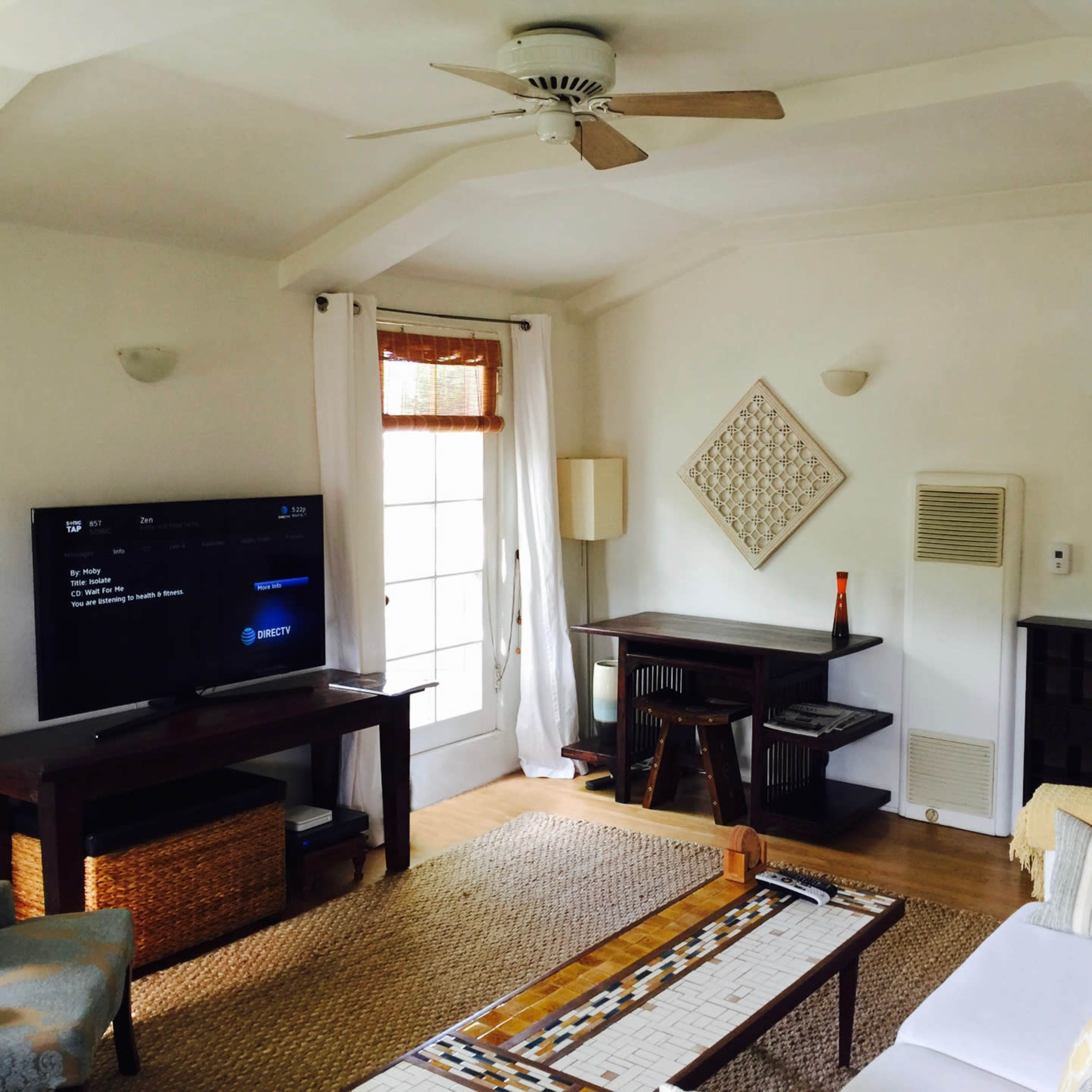 A living room features a television on a stand, a table with wooden chairs, a couch, and a door leading to an outdoor area.