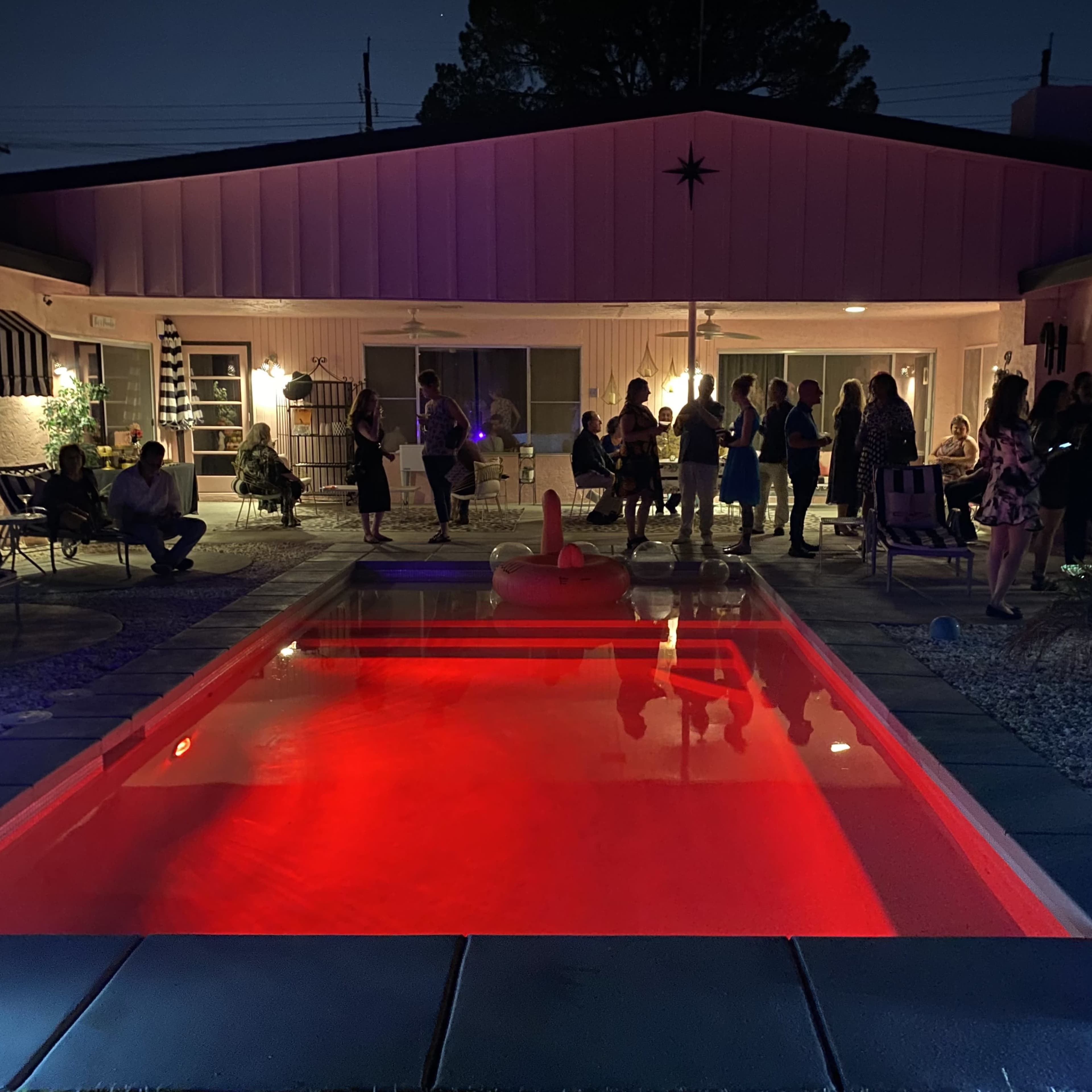 A group of people gathers around a brightly lit pool area in front of a house at night.