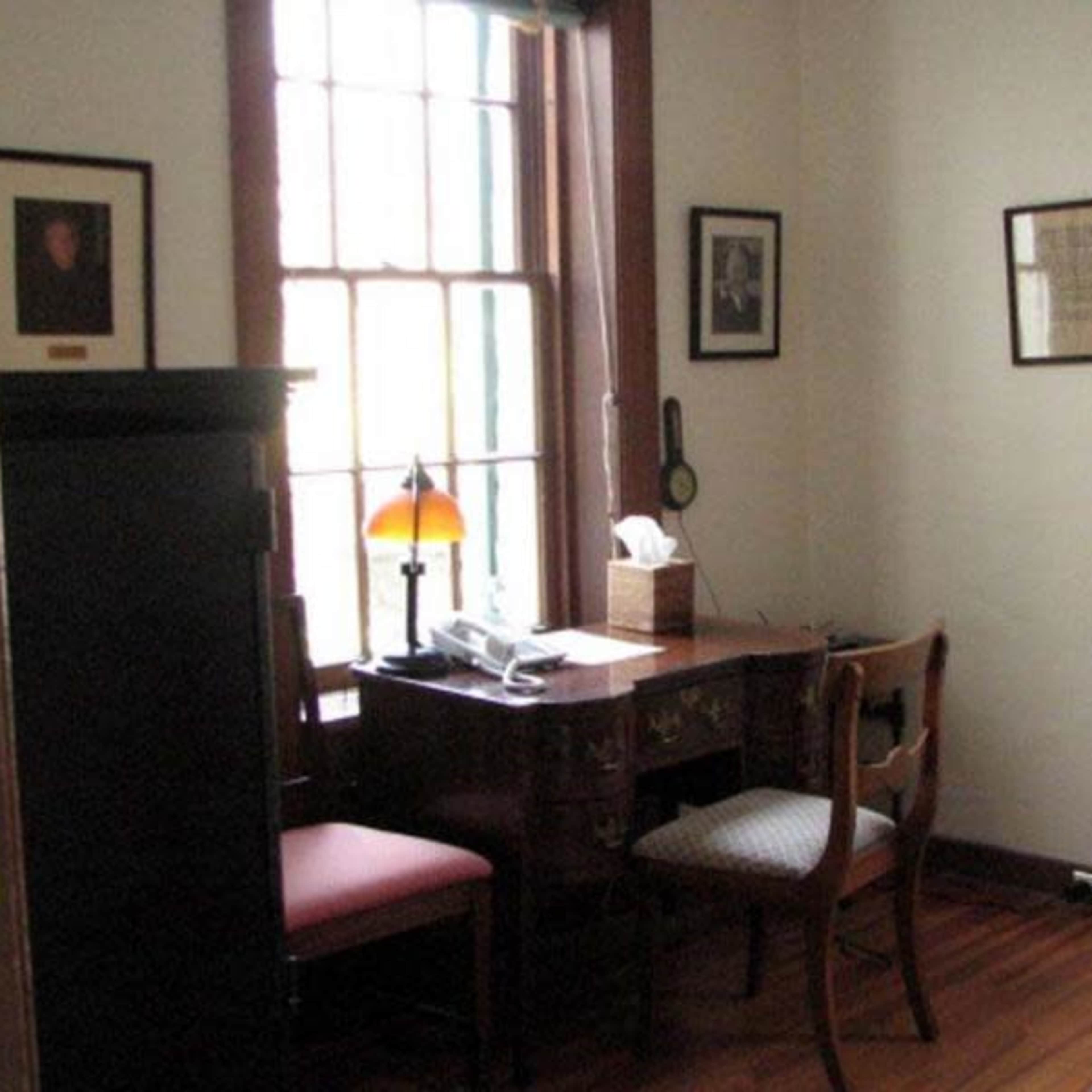 A small office space featuring a wooden desk with a lamp, a chair, and framed pictures on the walls.
