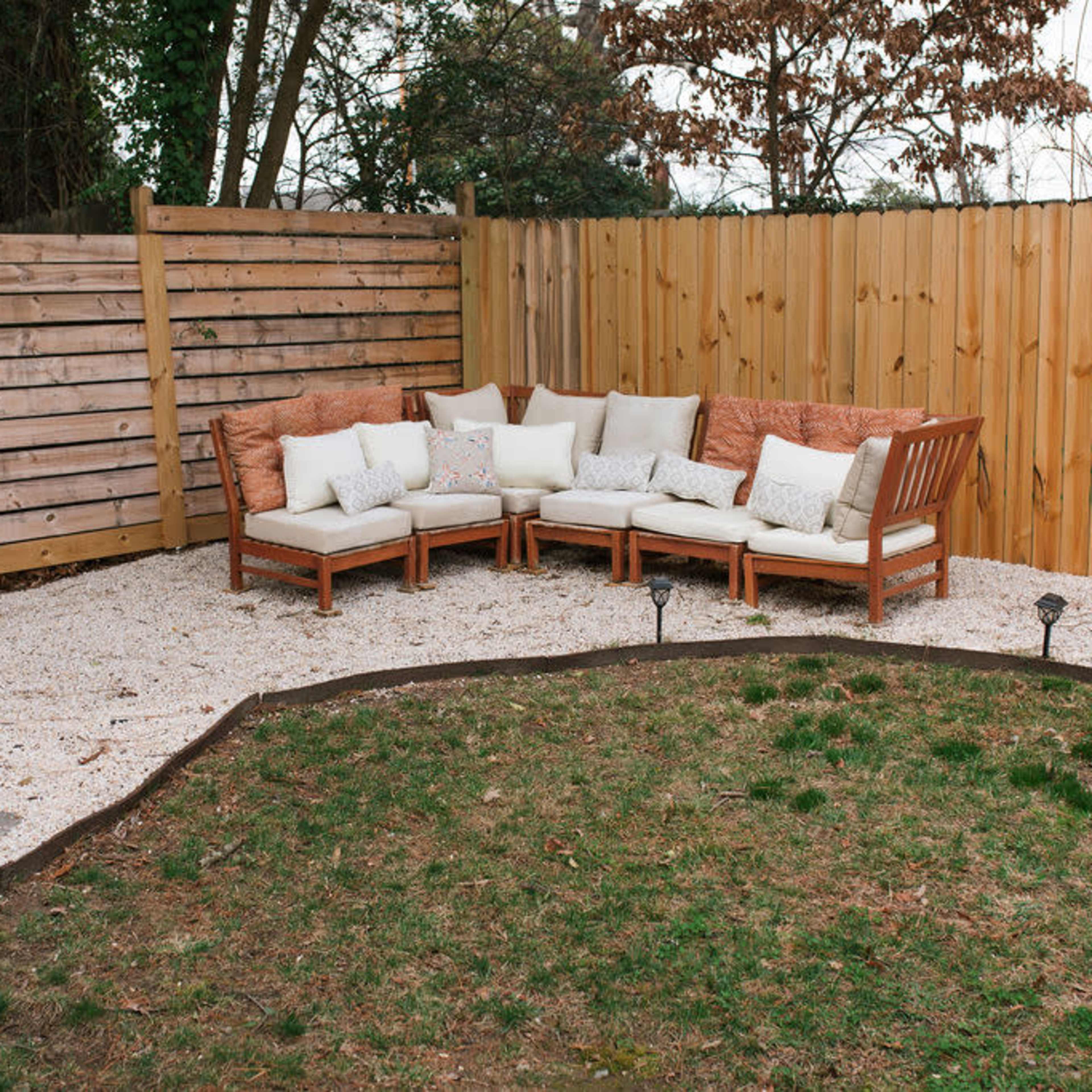 A wooden seating area with cushions is positioned on gravel in a fenced backyard, surrounded by grass and a decorative border.