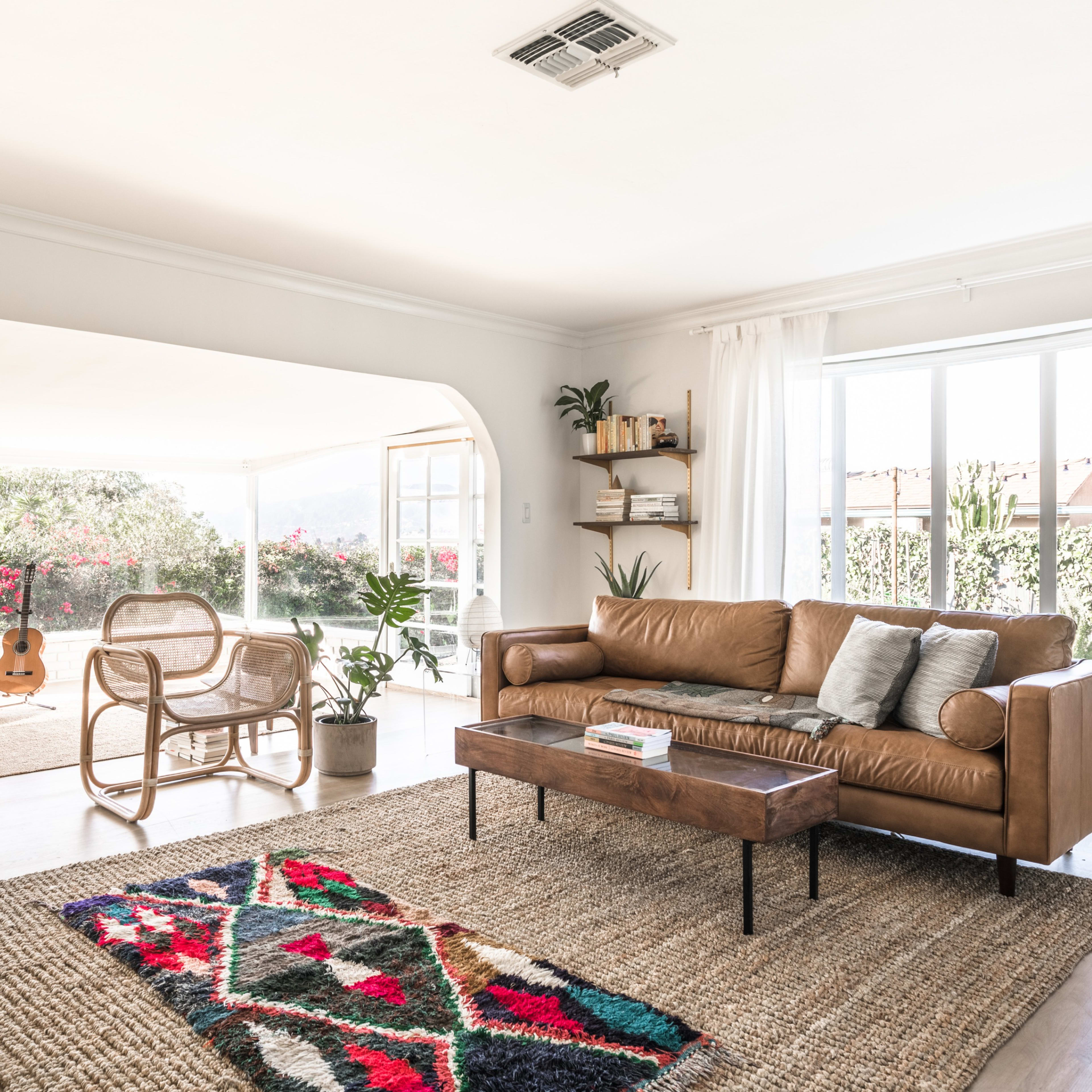 A spacious living room features a brown leather sofa, a wooden coffee table, and a guitar, with large windows offering a view of greenery and bright sunlight.
