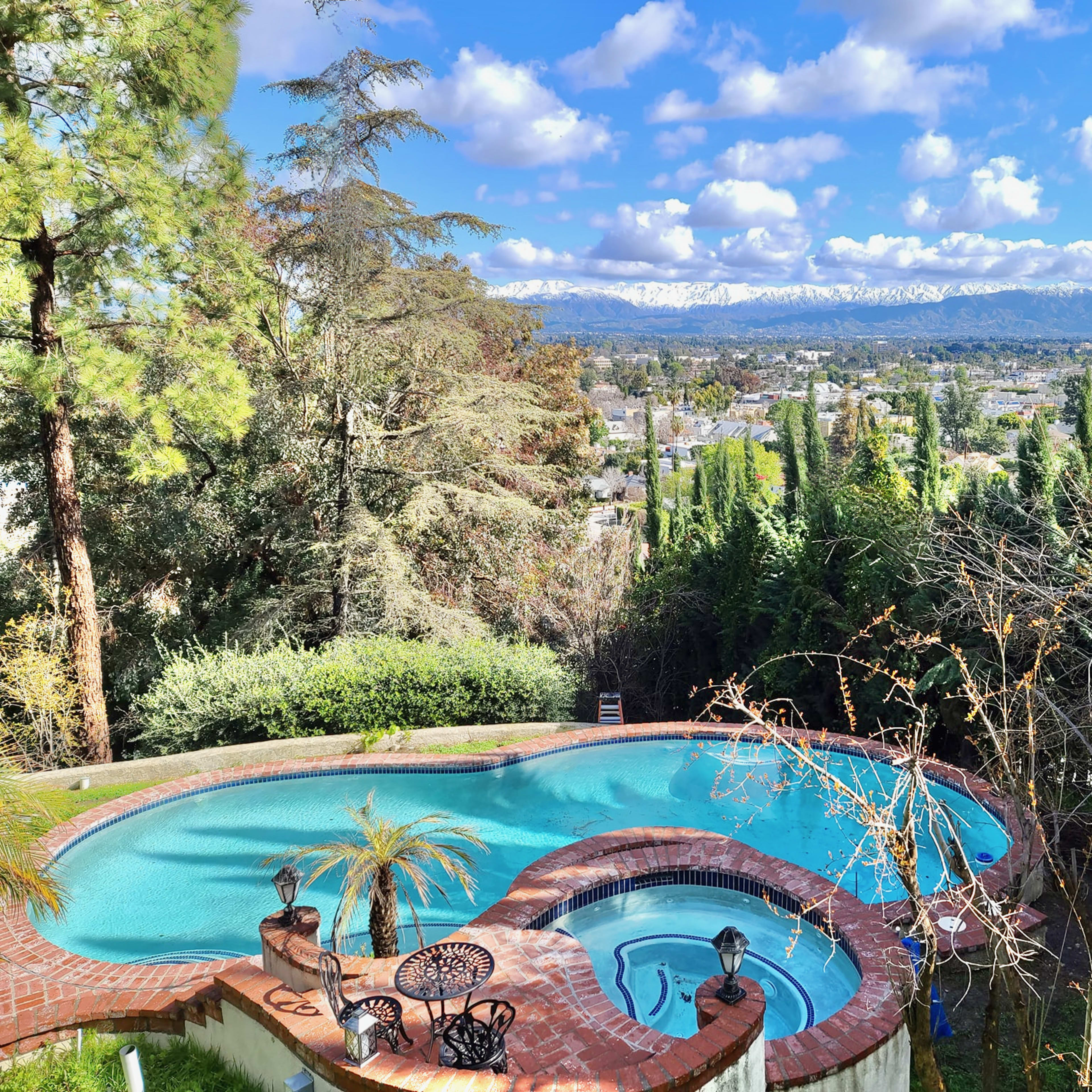 A swimming pool with a spa area, surrounded by trees and overlooking a mountainous landscape with a clear sky.