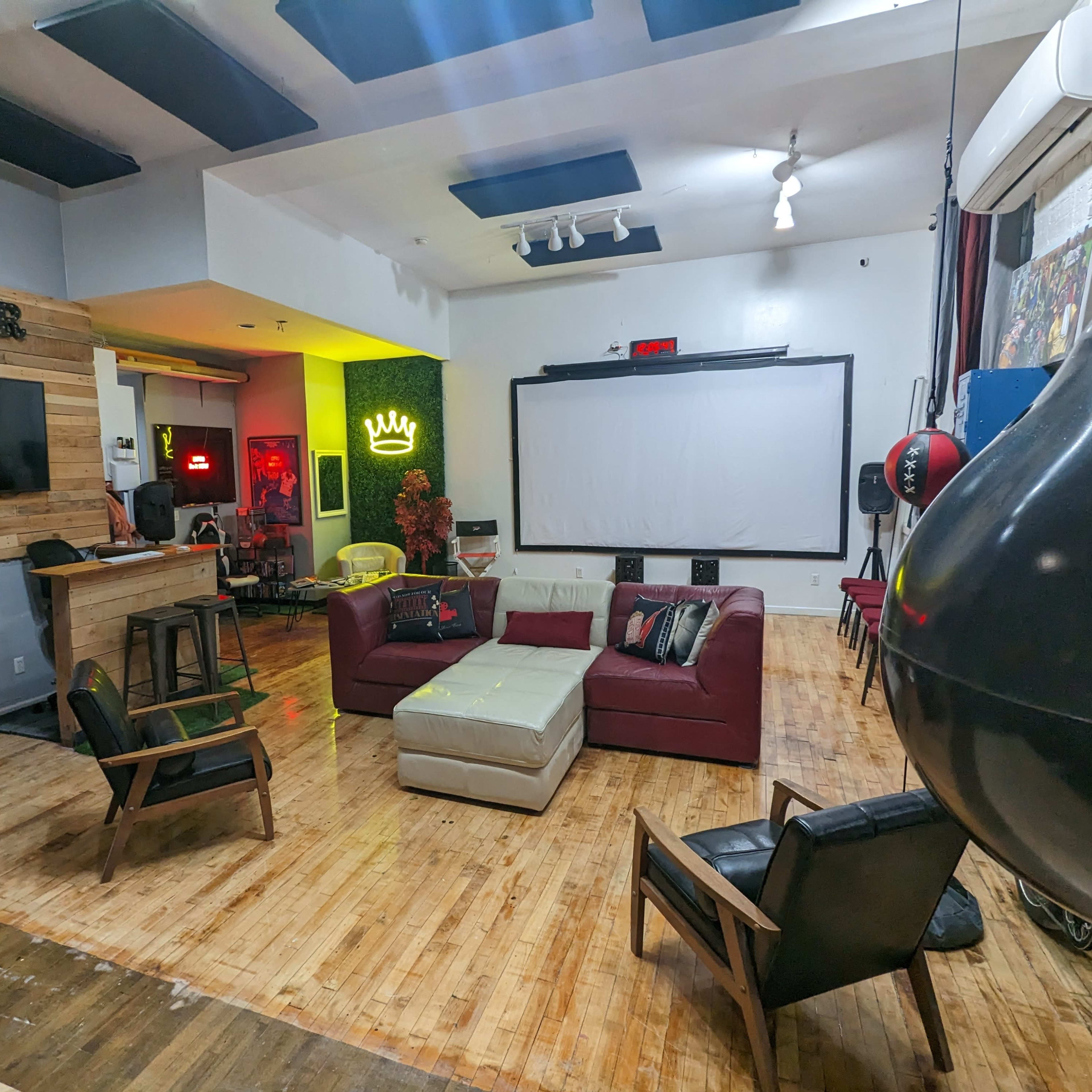A spacious lounge area with a red couch, a large projection screen, wooden flooring, and a bar on one side with various seating arrangements.