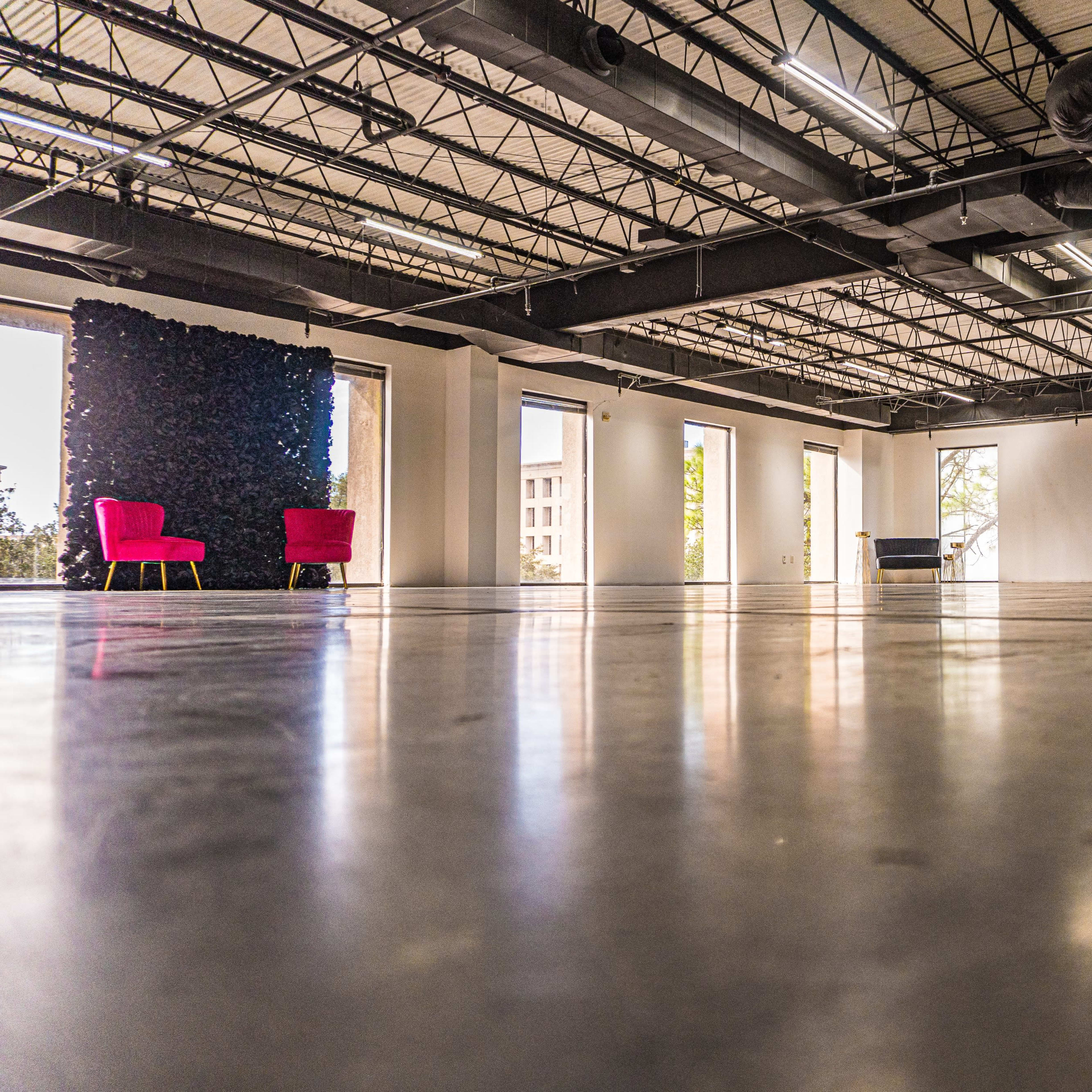 An empty modern space features polished concrete floors, large windows, and a pair of red chairs against a green wall.