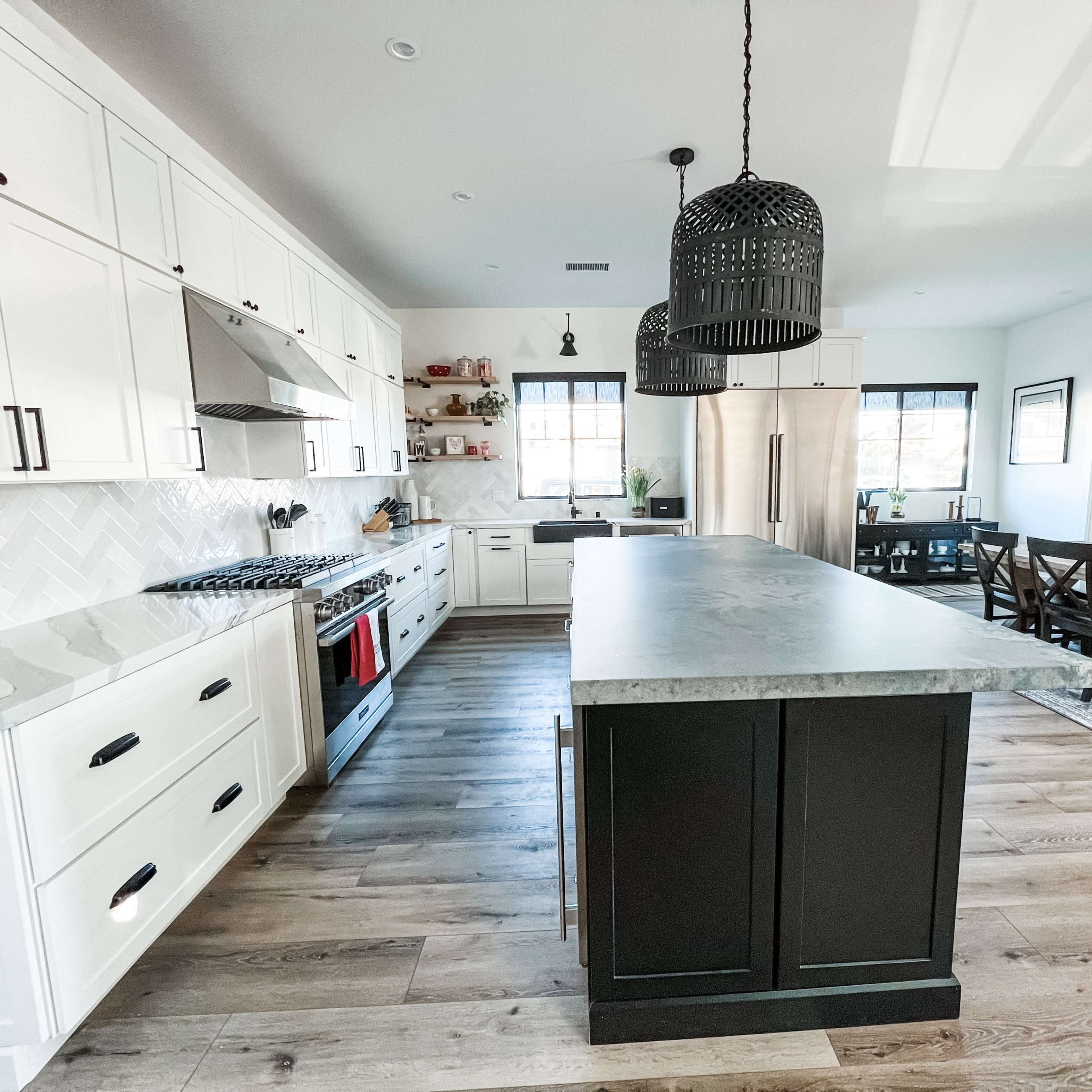 A modern kitchen features white cabinets, a large gray island, a gas range with a red accent, and a dining area with a wooden table.