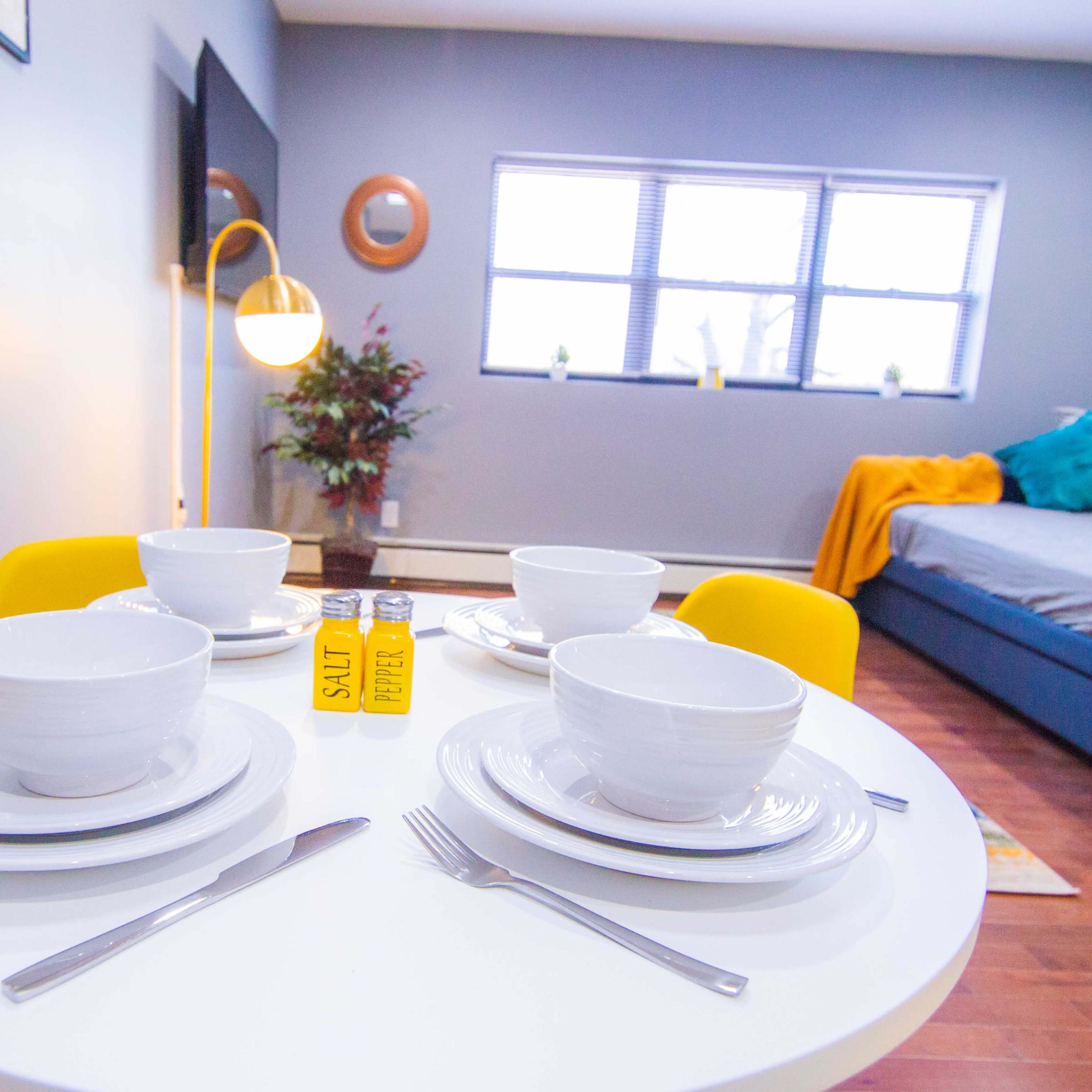 A small round table is set with four white bowls and utensils, with a yellow salt and pepper shaker in the center, in a room featuring a bed and decorative elements.