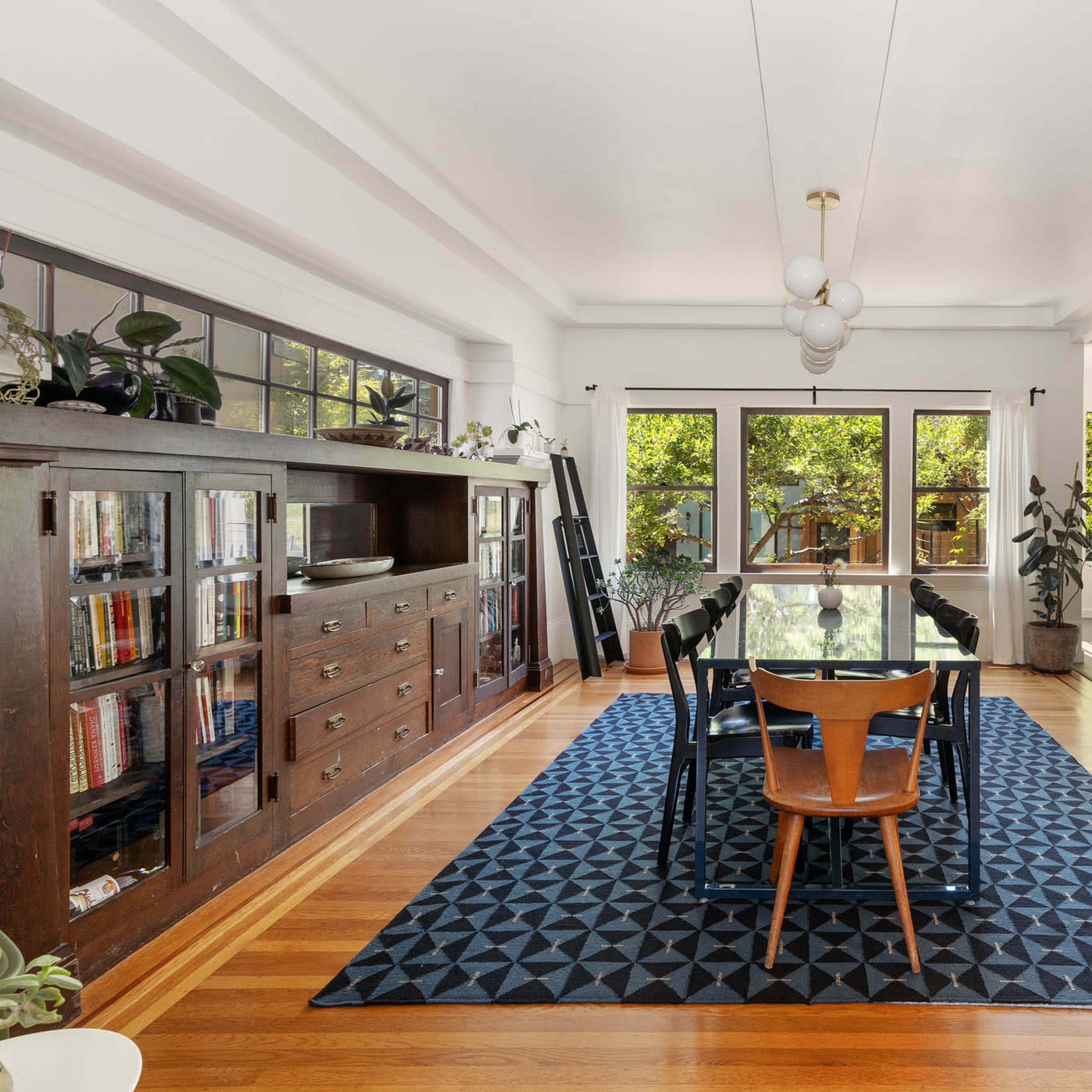 A dining area features a large rectangular table surrounded by chairs, with large windows overlooking a green outdoor space and wooden flooring.