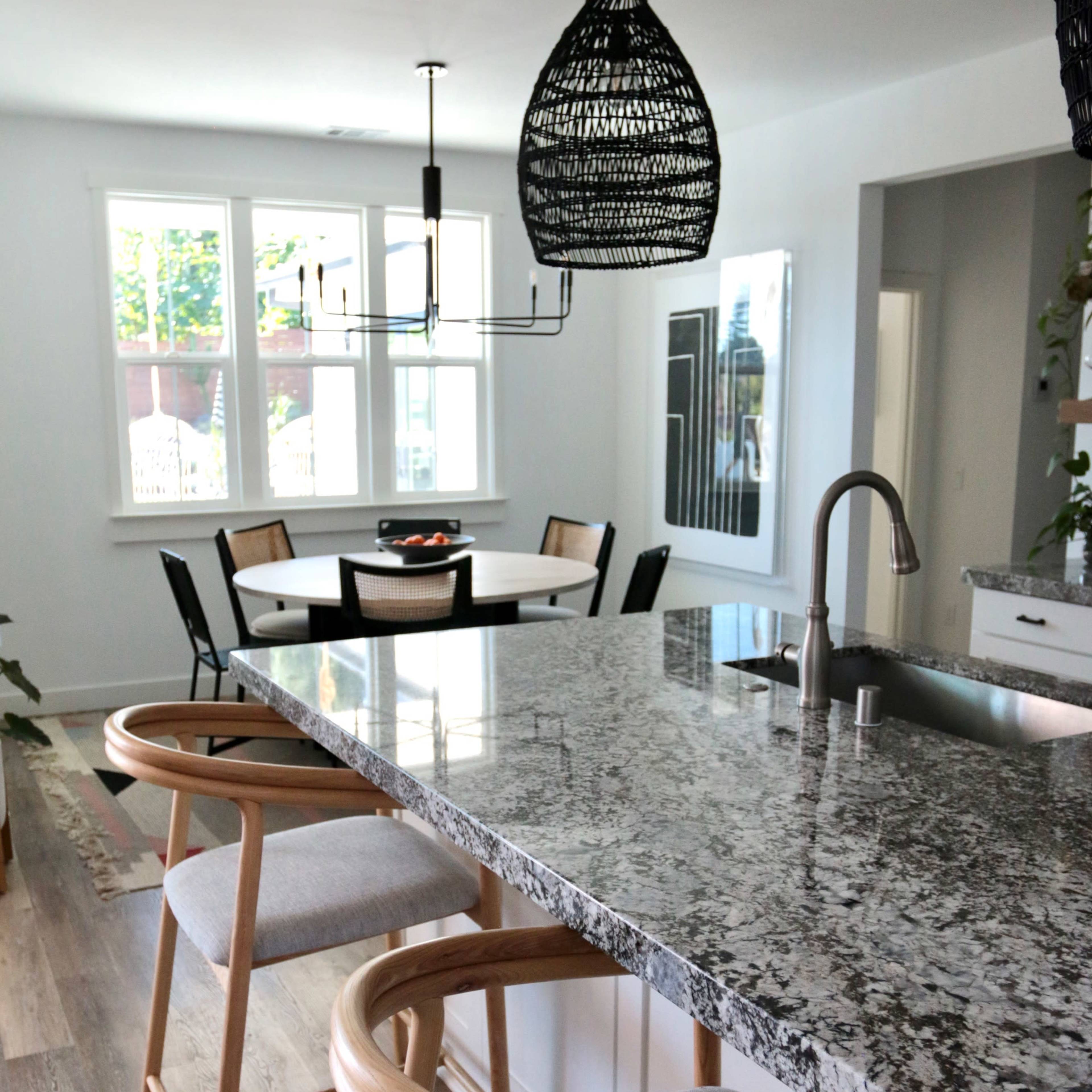A modern kitchen features a granite countertop with two wooden bar stools, pendant lights above, and a dining area visible through a large window.