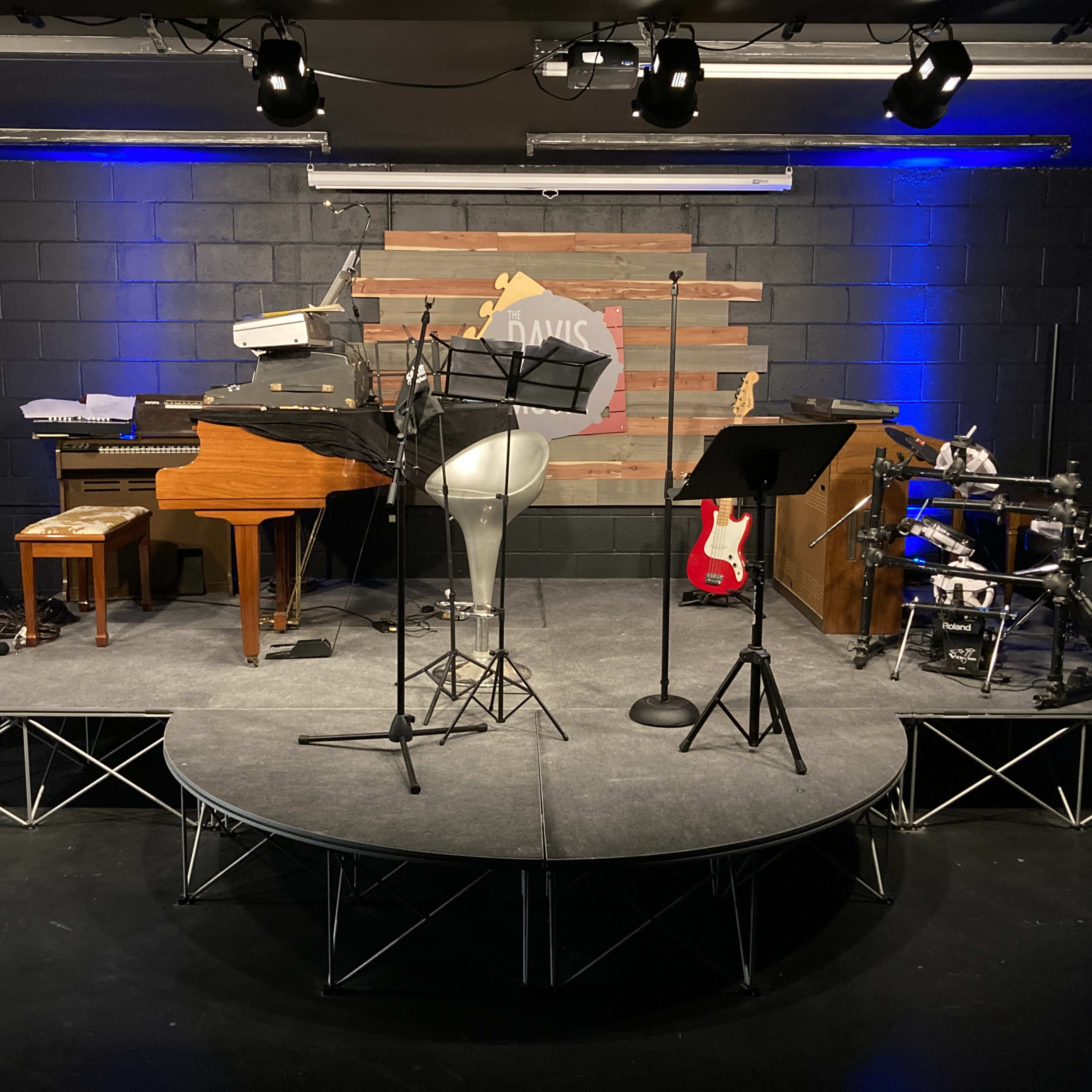 A small stage set up with a grand piano, a microphone stand, musical instruments, and music stands against a backdrop of wooden panels.