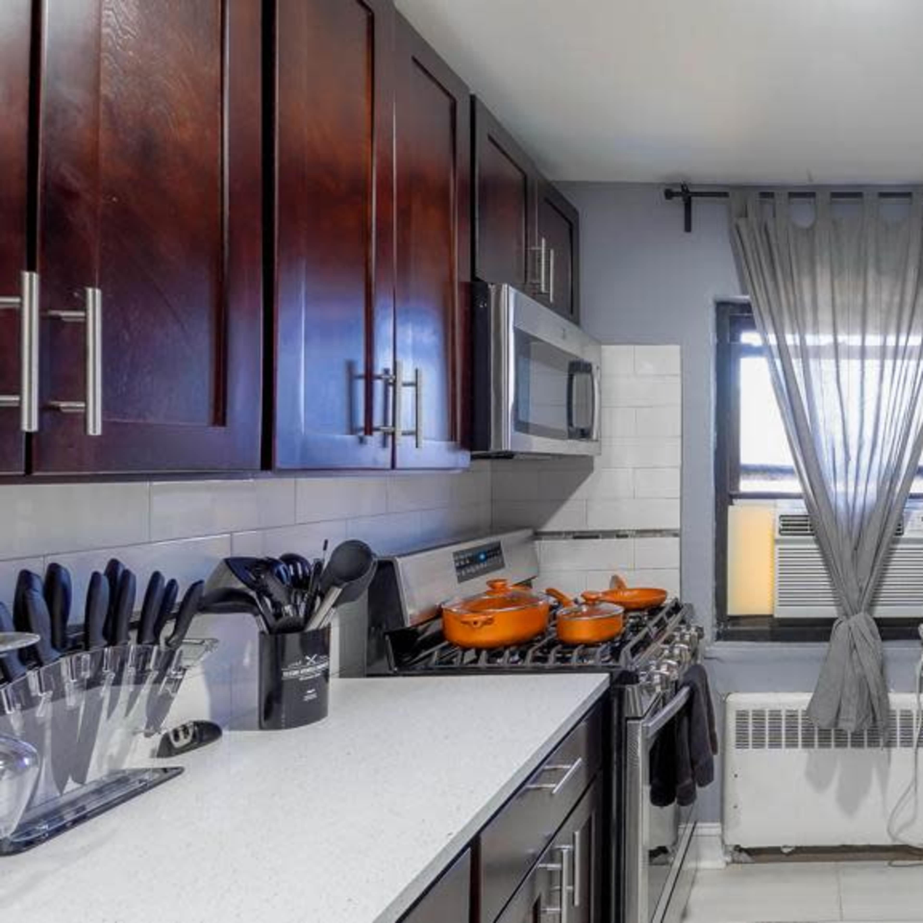 A modern kitchen with dark wooden cabinets, a window with light curtains, and a countertop with cooking utensils and orange pots.