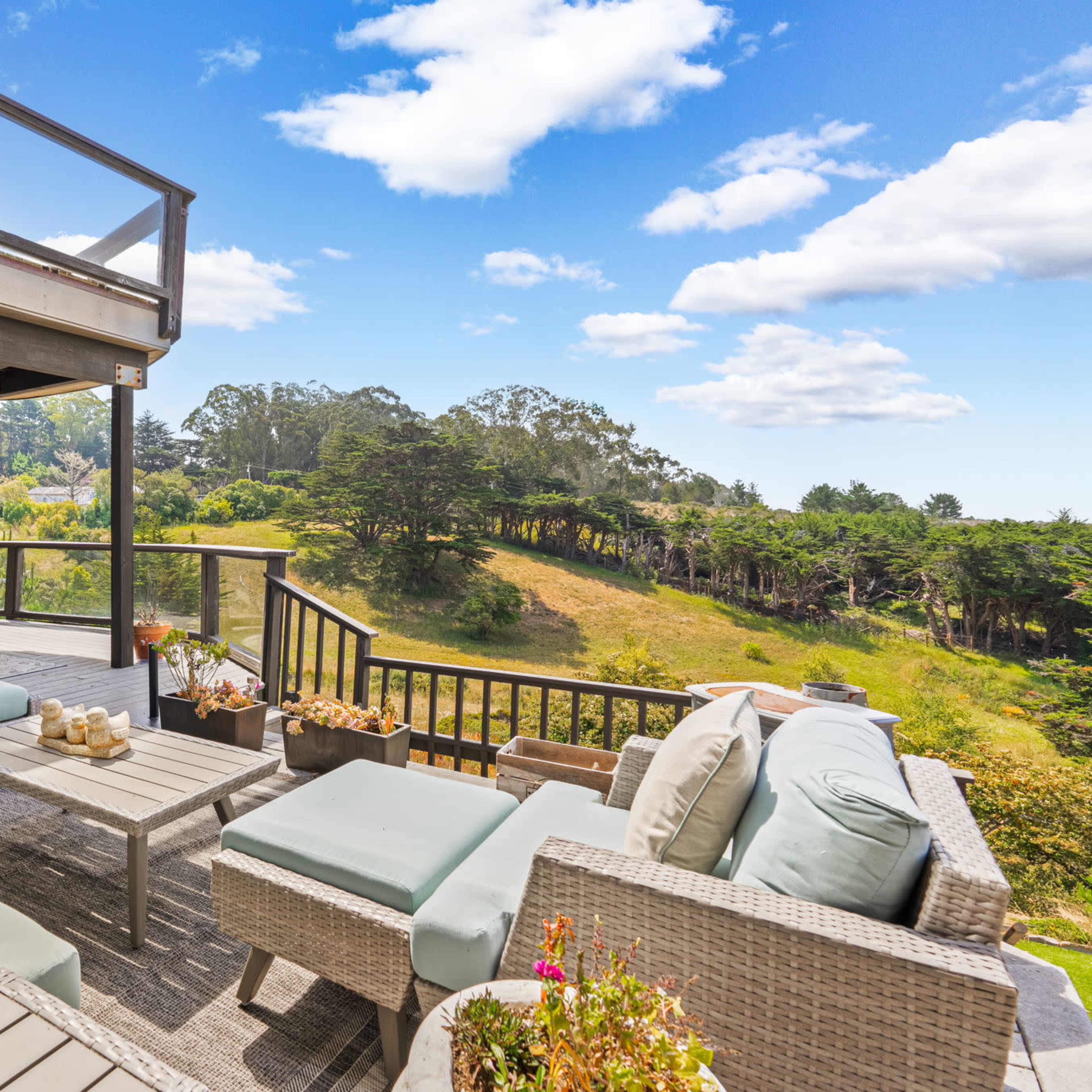 A spacious outdoor terrace features comfortable seating and a panoramic view of a green hillside under a blue sky with scattered clouds.