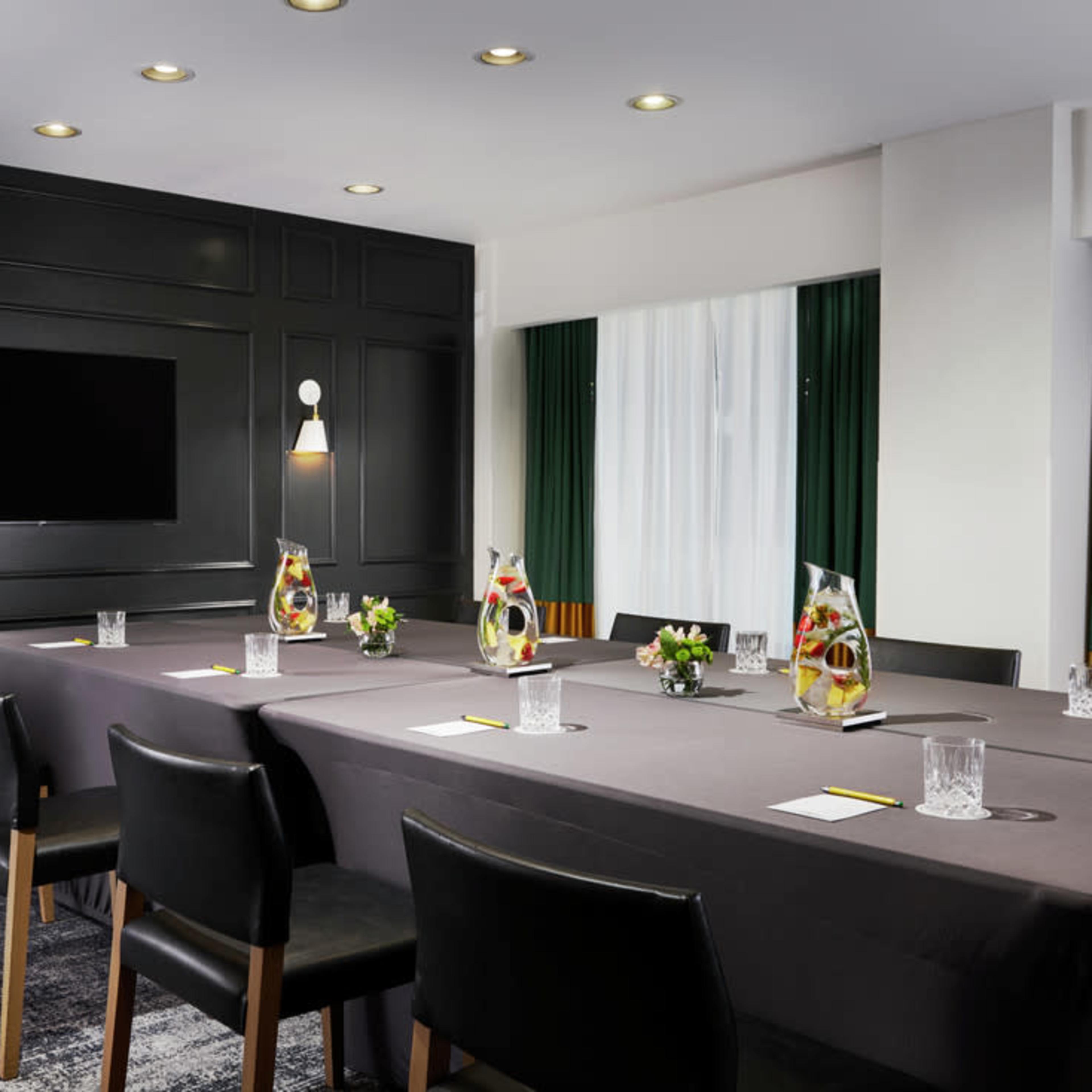 A conference room features a long table set with water pitchers, glasses, and floral centerpieces, surrounded by black chairs and illuminated by ceiling lights.