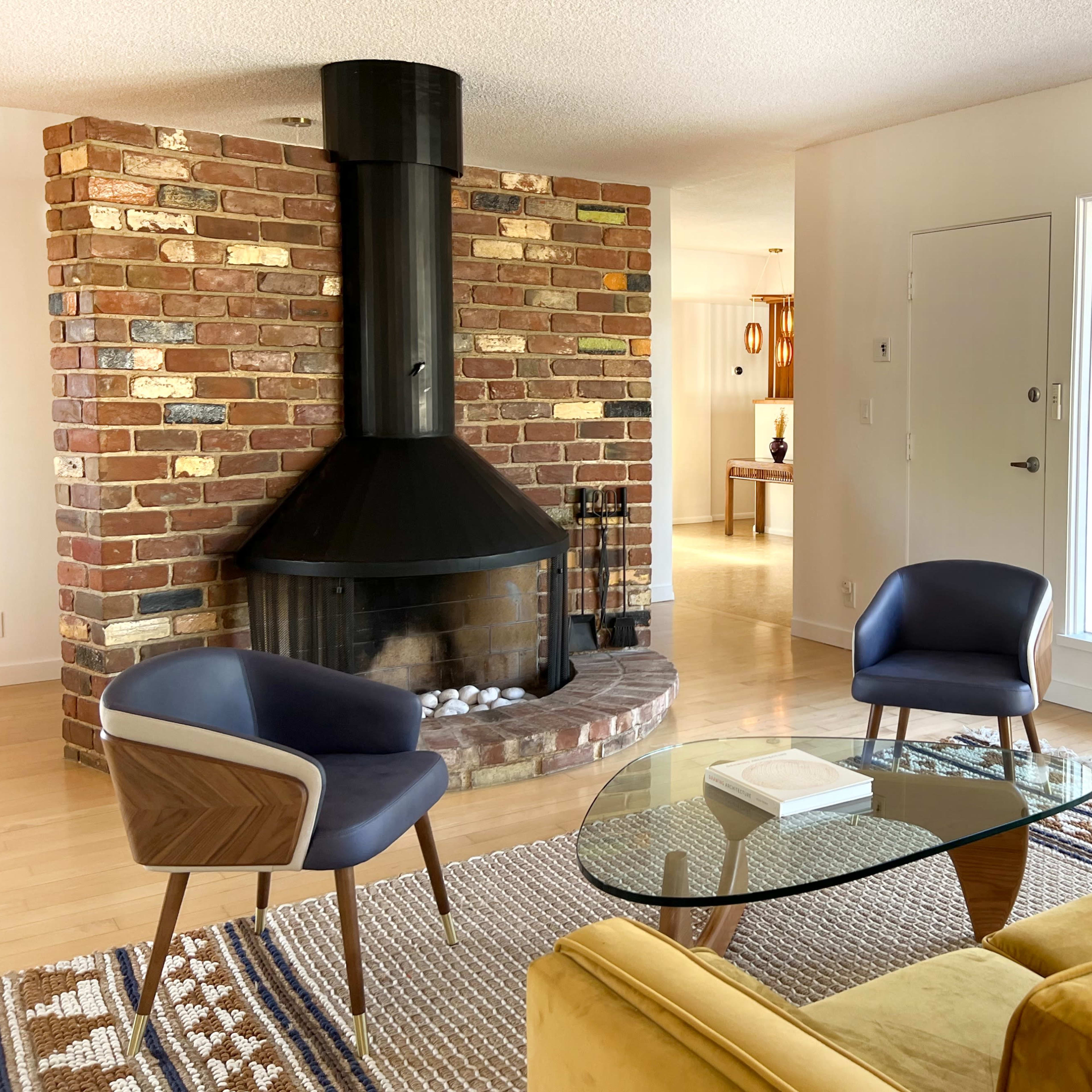 A stylish living room with a black chimney-style fireplace, two blue armchairs, a glass coffee table, and a yellow sofa, surrounded by brick walls and large windows.
