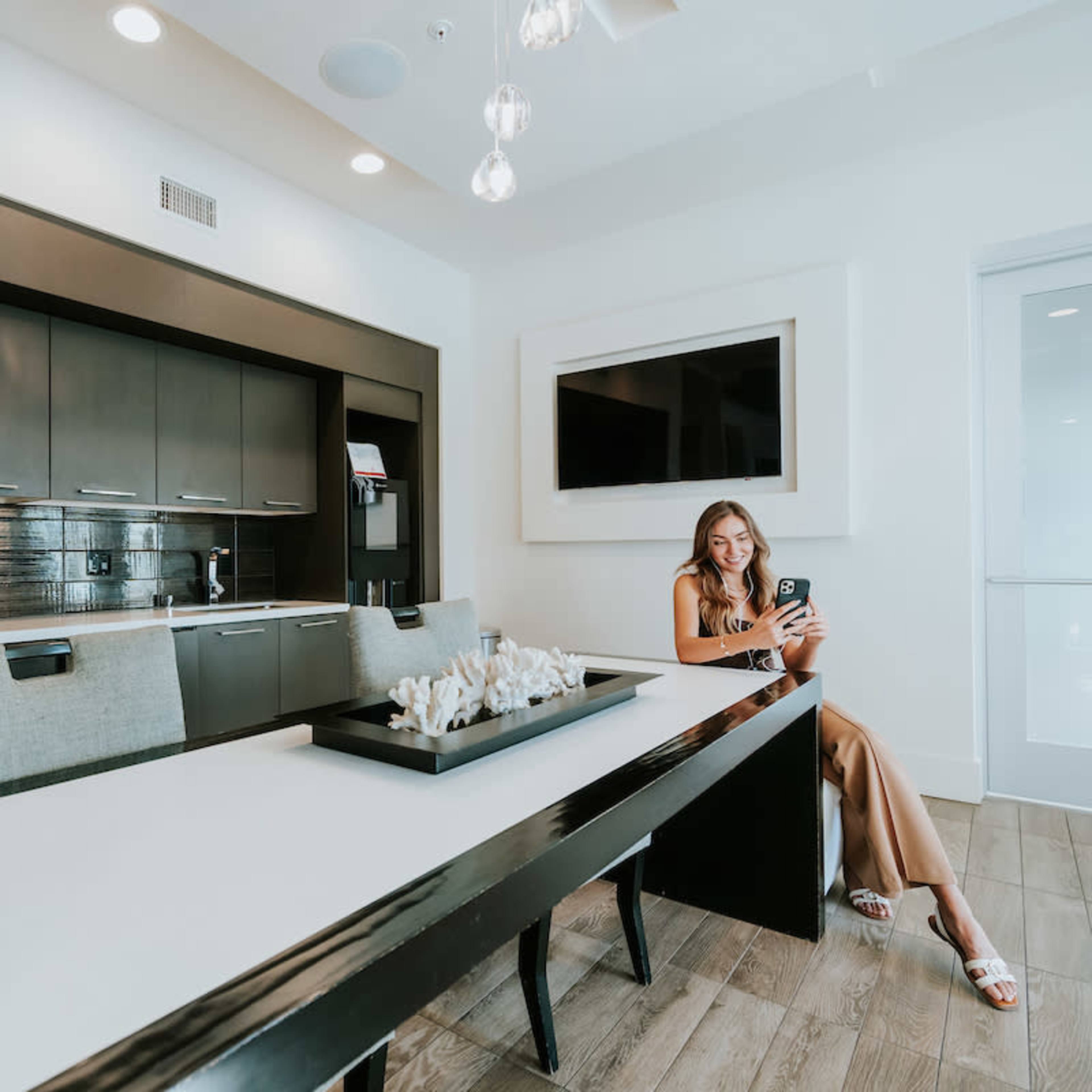 A woman sits at a long dining table in a modern kitchen with dark cabinetry and a large wall-mounted television.