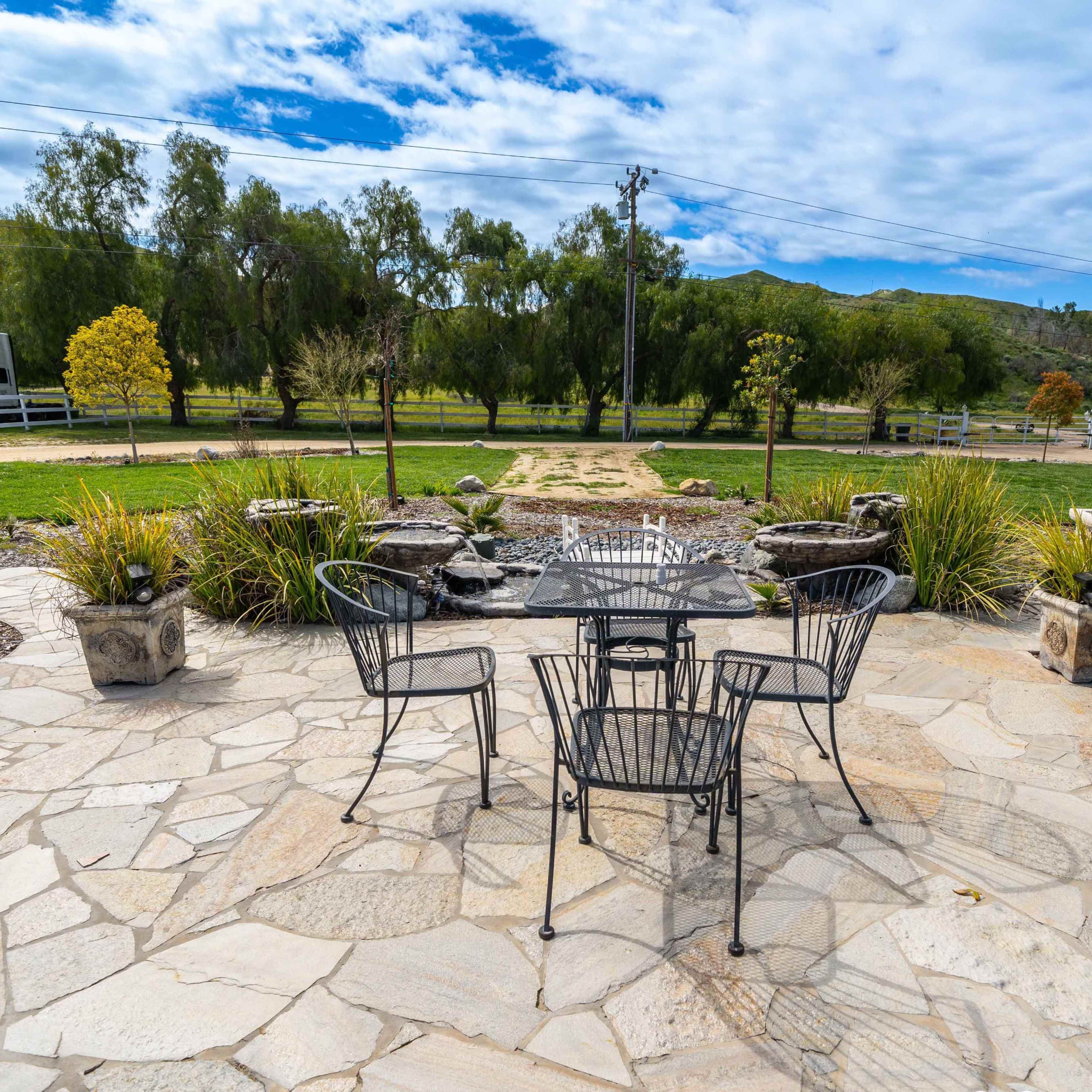 A stone patio with a round table and four black chairs overlooks a green landscape with trees and a white fence in the background.