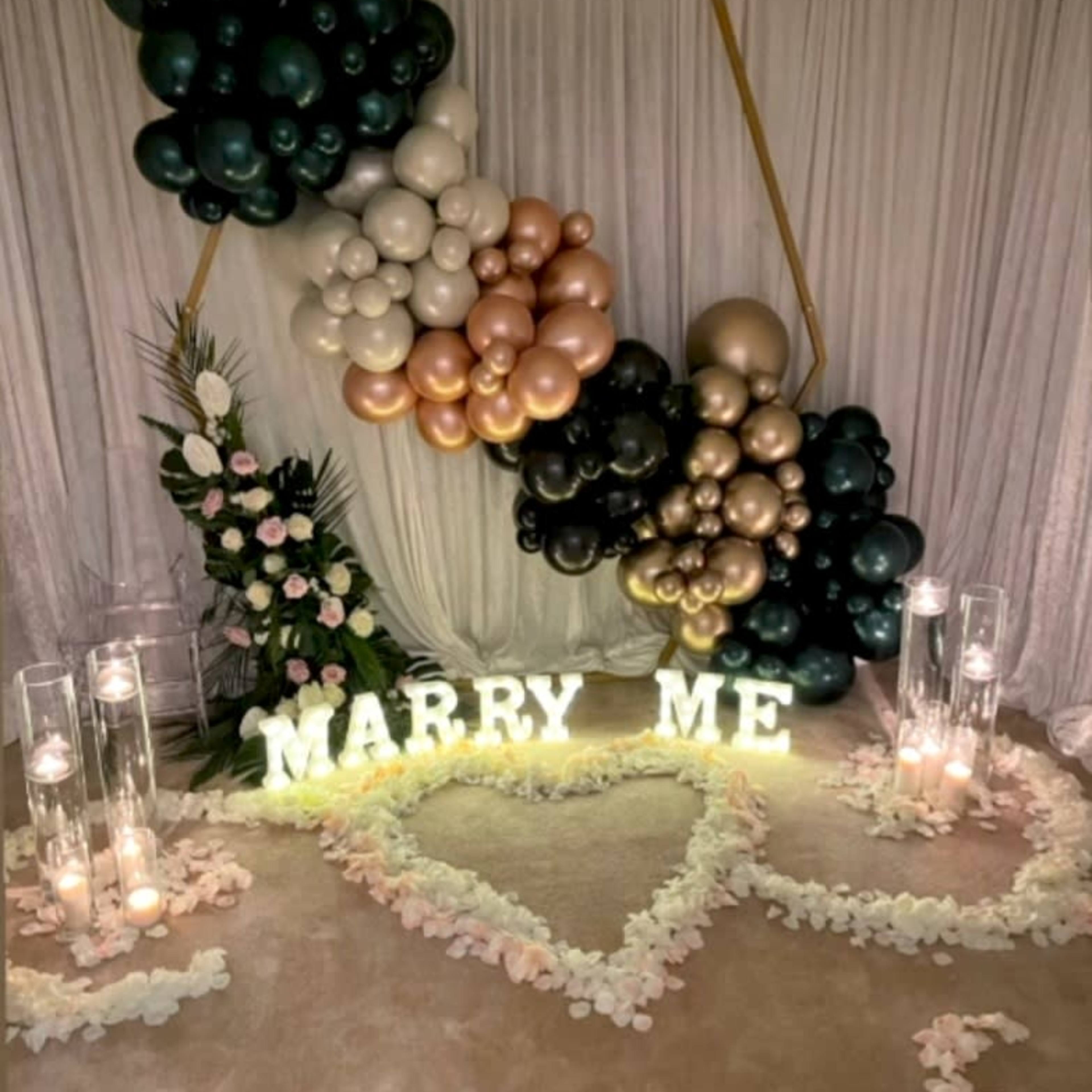 A decorative setup for a proposal, featuring a heart-shaped arrangement of flower petals, illuminated letters spelling "MARRY ME," and a backdrop of balloons in various colors.