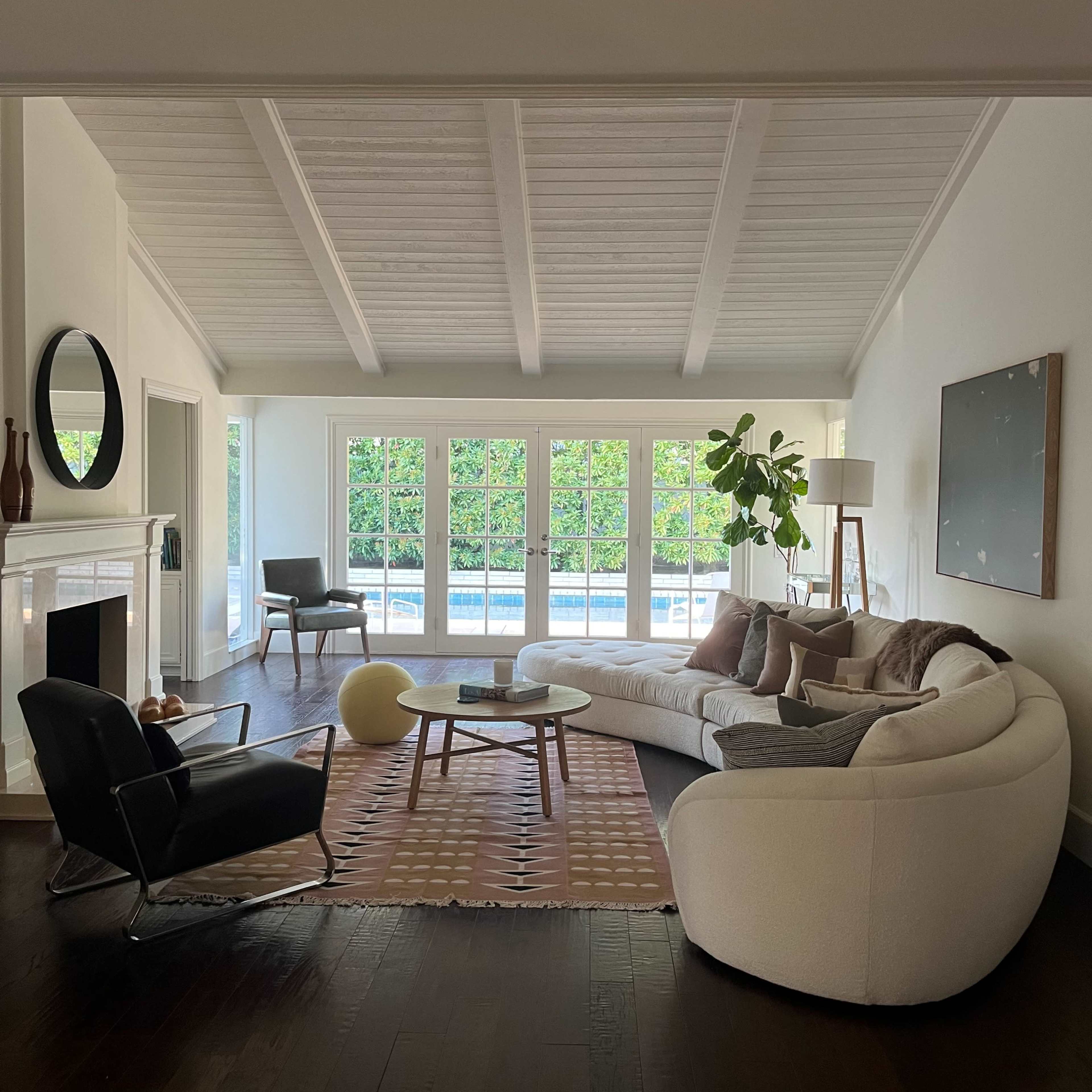 A modern living room features a curved white sofa, a black armchair, and large windows overlooking a pool, with a cozy rug and decorative plants.