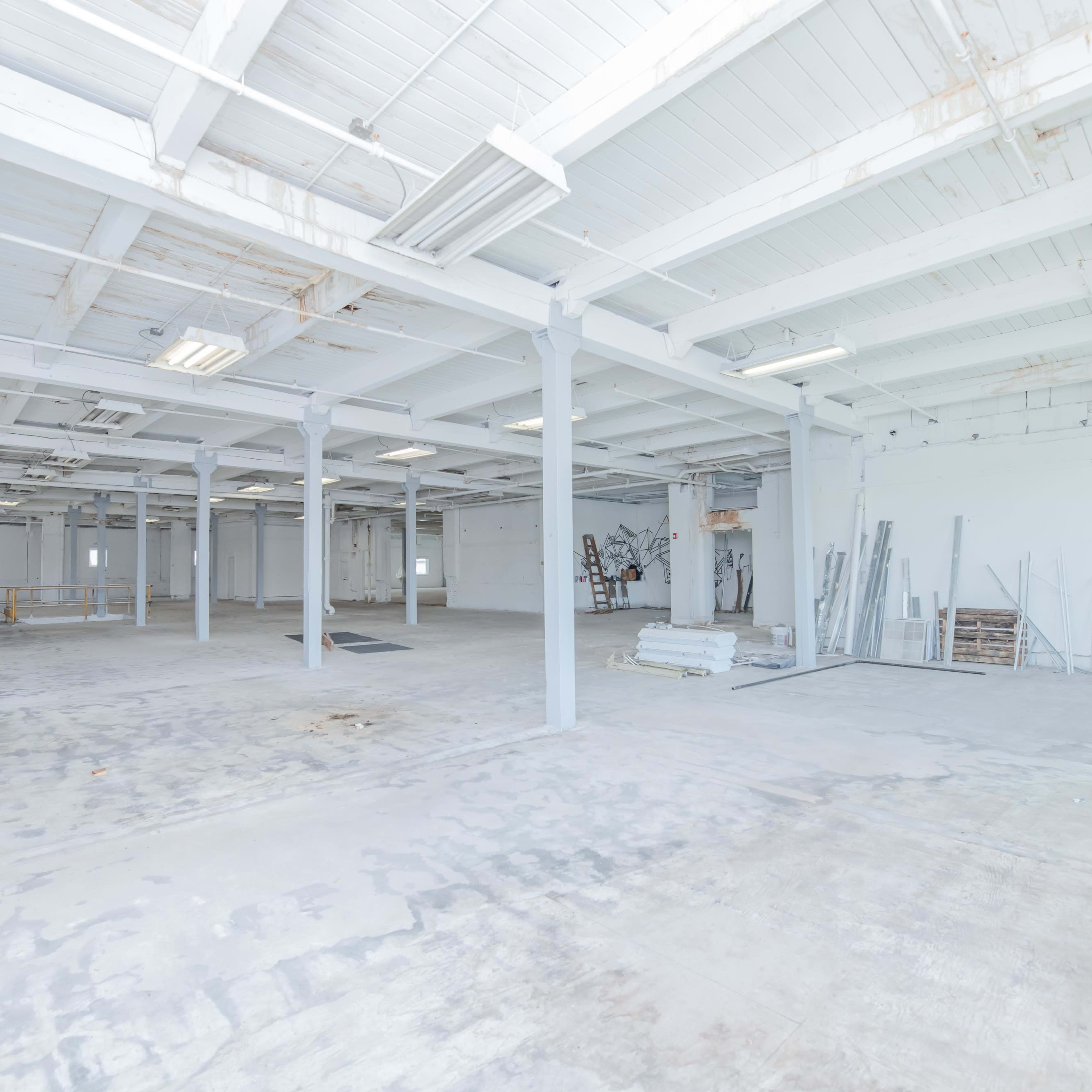 A large, empty industrial space with concrete floors and white walls, featuring exposed beams and minimal furnishings.