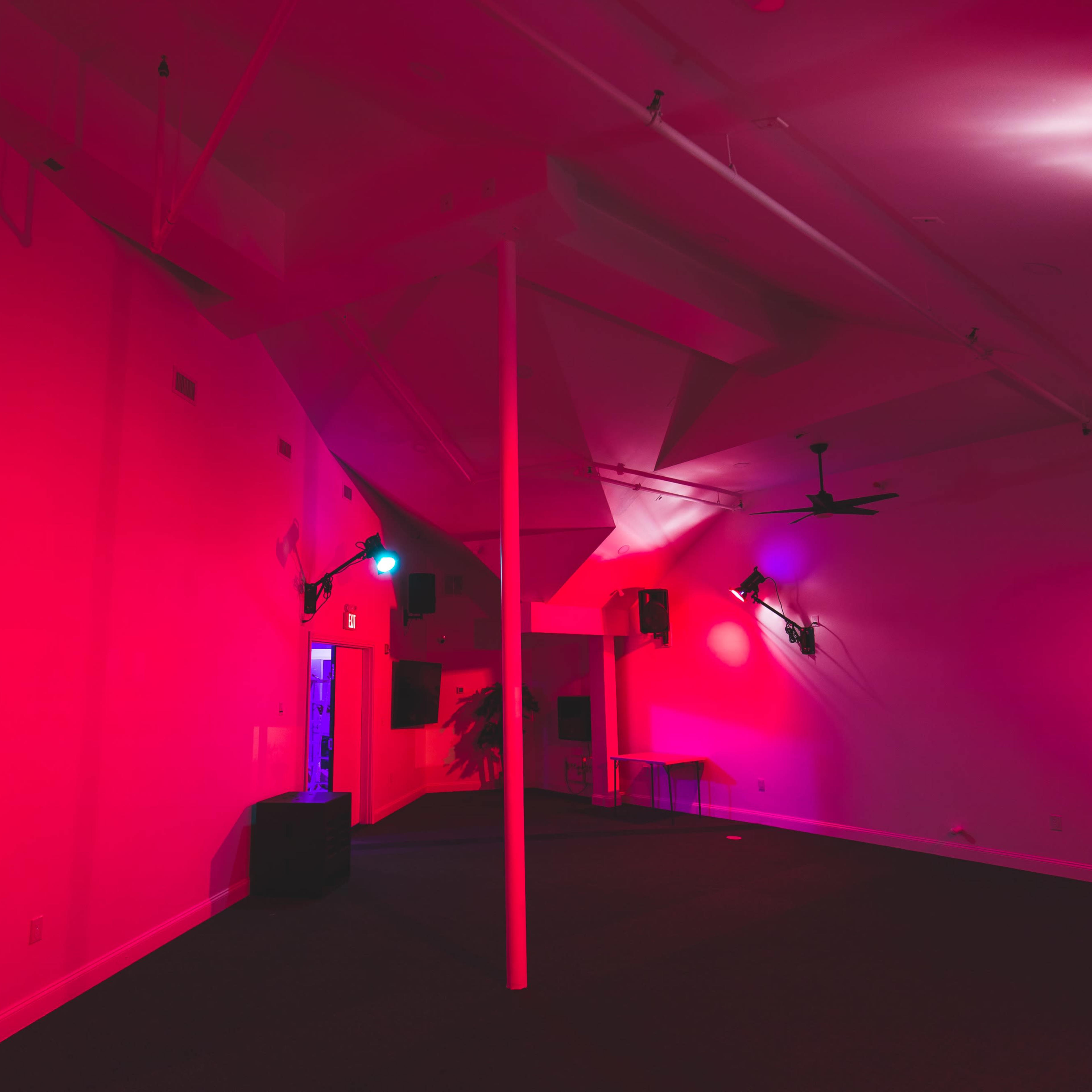 An empty room with a central pole, colored lighting in shades of pink and blue, and various mounted speakers on the walls.